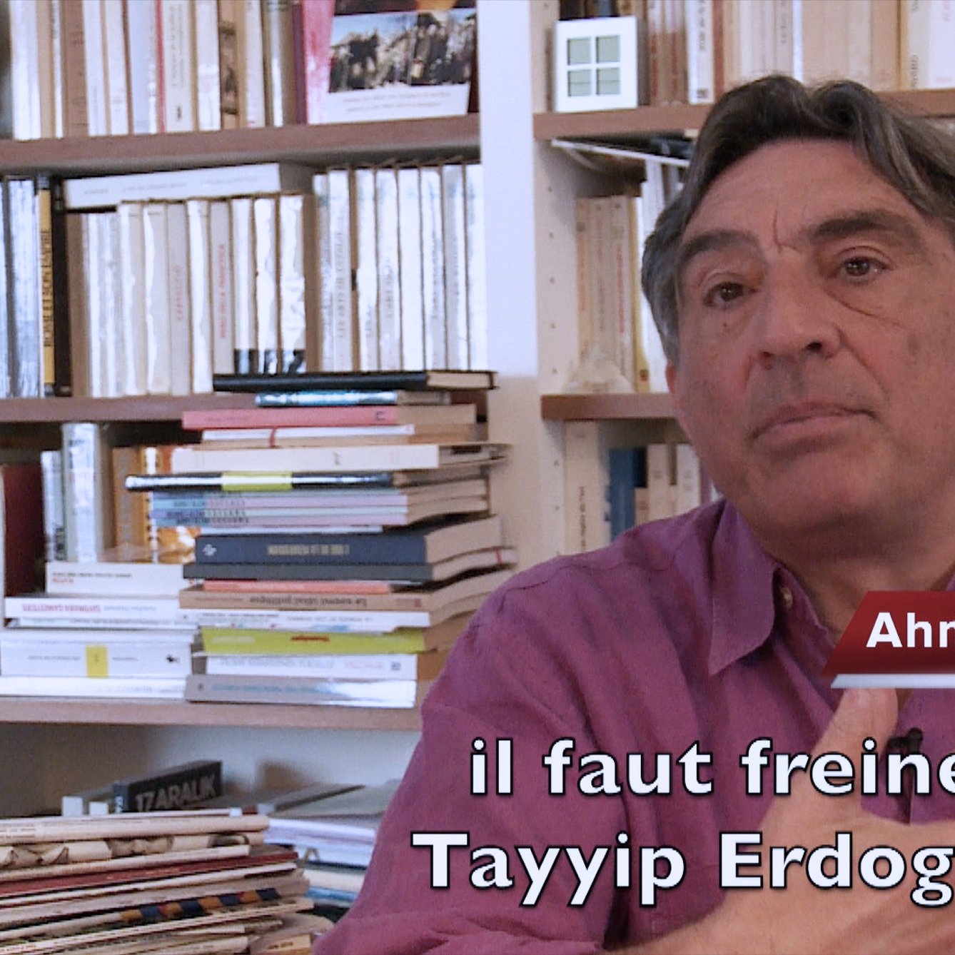 L'image montre un homme assis dans un environnement intérieur, entouré de livres et de dossiers sur des étagères. Il semble s'adresser à un interlocuteur ou à une caméra. L'homme est identifié comme Ahmet Insel, un professeur, et il exprime une opinion sur la nécessité de freiner Tayyip Erdogan. Il porte une chemise de couleur mauve et utilise des gestes pour appuyer ses propos. Les livres en arrière-plan suggèrent un cadre intellectuel.