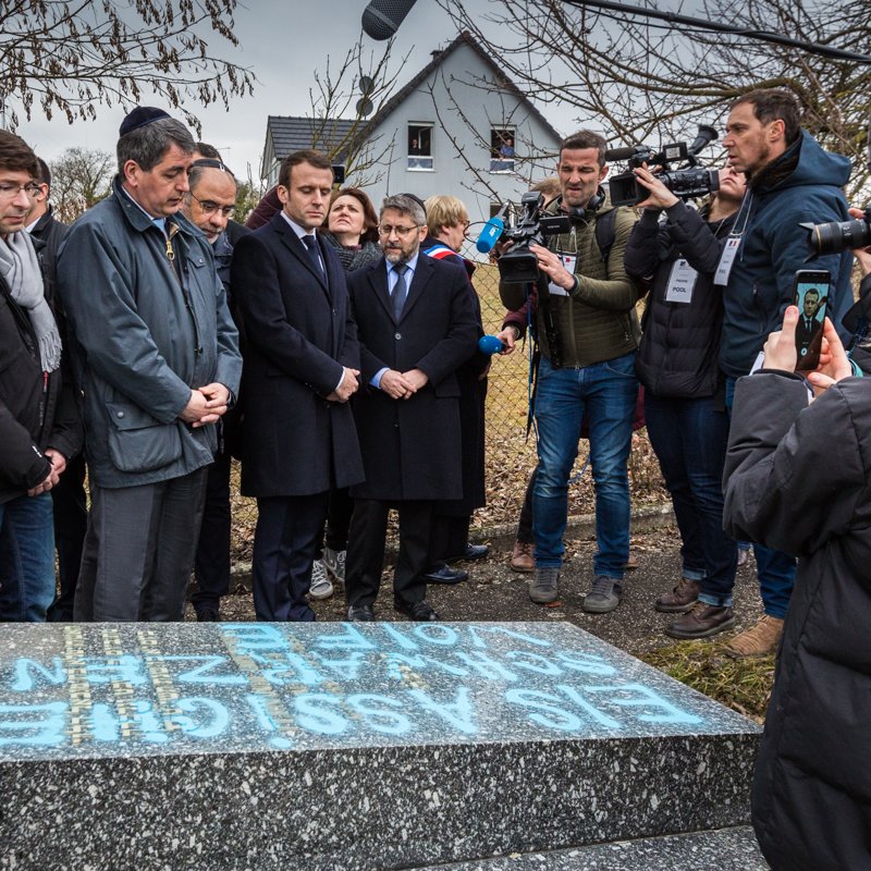 L'image montre un groupe de personnes rassemblées autour d'une stèle ou d'une tombe. Parmi elles, on peut distinguer des individus en tenue formelle, semblant assister à une cérémonie hommage. Un enfant tient un téléphone pour filmer ou prendre une photo. Des journalistes, avec des caméras et des micros, sont également présents, suggérant que l'événement est médiatisé. En arrière-plan, on aperçoit une maison et des arbres, indiquant un cadre extérieur. 