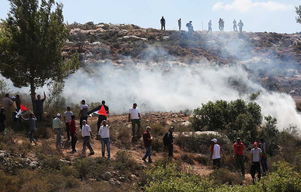 تظهر الصورة مجموعة من الأشخاص في منطقة مفتوحة، بعضهم يحمل الأعلام الفلسطينية. يبدو أن الاحتجاجات أو المظاهرات جارية، حيث تتصاعد سحب الدخان في الخلفية. يتمركز عدد من الأشخاص على تلة، مما يشير إلى وجود توتر أو حدث مهم في المكان. الطبيعة المحيطة تتميز بالأشجار والأراضي الوعرة.