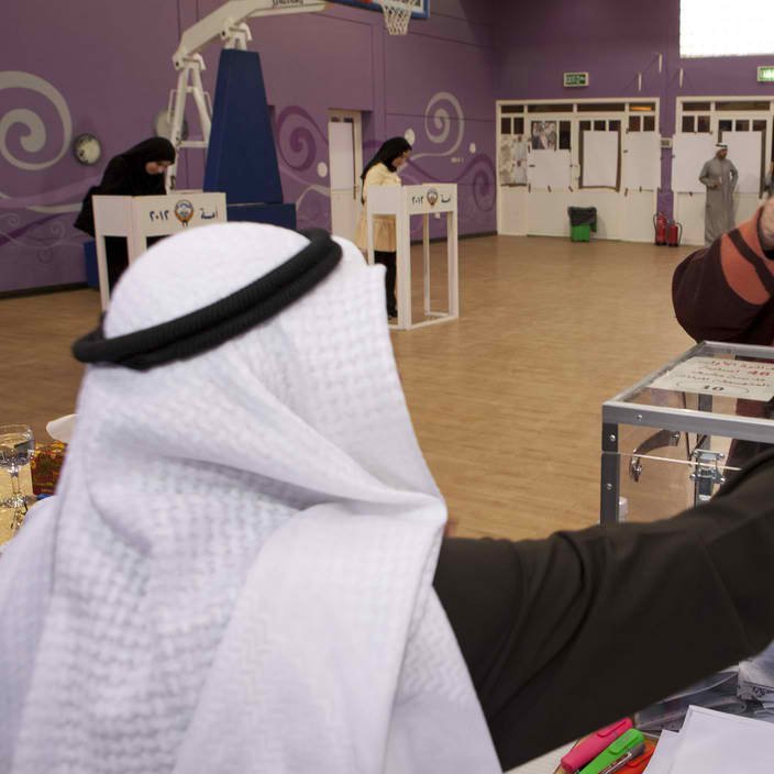 L'image montre une scène d'élection dans un environnement intérieur. Au premier plan, un homme portant un kandora et un ghutrah se tient derrière une urne électorale. Il regarde une femme voilée qui tend un bulletin de vote, le visage partiellement caché par un niqab. En arrière-plan, on peut voir d'autres personnes et des isoloirs, indiquant que le processus de vote est en cours dans cet espace, qui semble être une salle de sport ou un centre communautaire. La décoration du mur est colorée, ajoutant une atmosphère dynamique à l'événement.