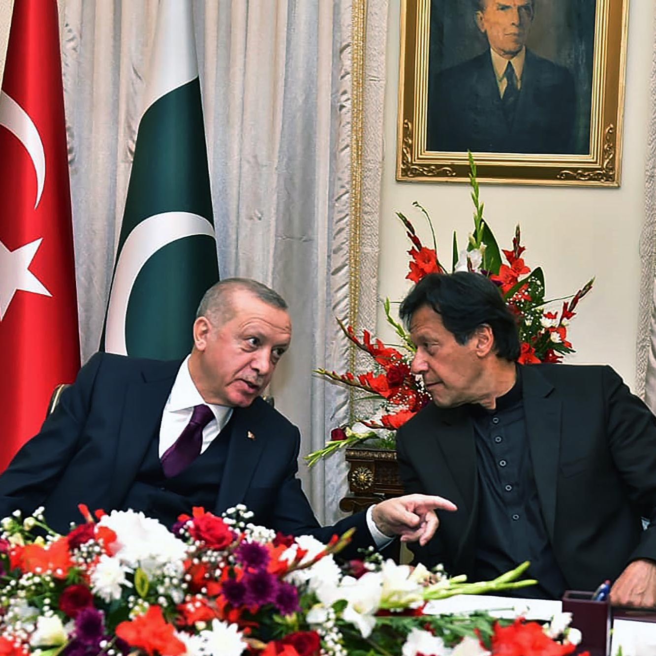 L'image montre deux hommes en conversation, assis à une table décorée de fleurs. En arrière-plan, on peut voir des drapeaux turc et pakistanais, ainsi qu'un portrait accroché au mur. Les hommes semblent échanger des idées de manière animée, et l'atmosphère semble conviviale.