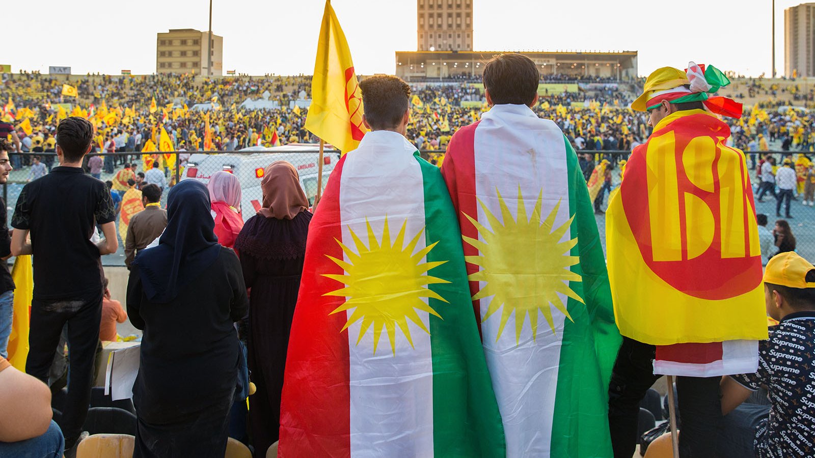 L'image montre un événement festif dans un stade rempli de supporters. Au premier plan, deux personnes portent des drapeaux, l'un étant le drapeau kurde avec les étoiles et l'autre avec un symbole qui semble représenter un mouvement ou un parti. Le public derrière eux est également vêtu de couleurs vives, principalement du jaune, suggérant une ambiance célébratoire. La scène évoque un fort sentiment d'unité et de fierté culturelle.