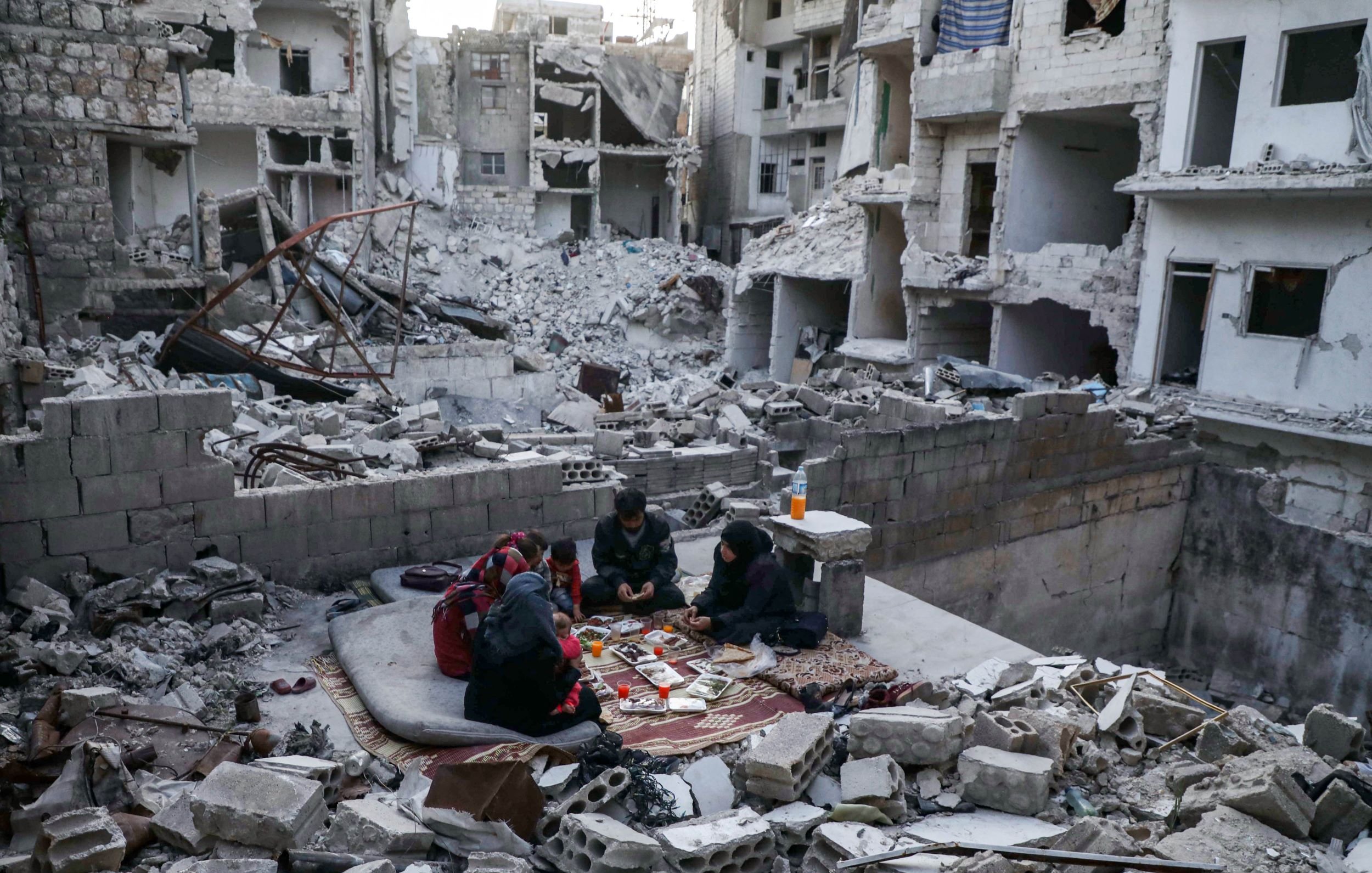 تصویر نشان‌دهنده‌ی یک منطقه ویران در یک شهر است که آثار تخریب شدید در آن مشهود است. ساختمان‌ها به شدت آسیب دیده و دیوارها و سقف‌ها خراب شده‌اند. در وسط این ویرانی، گروهی از افراد نشسته‌اند و روی زمین، میان خرابه‌ها، در حال بازی یا سرگرمی هستند. فضای کلی تصویر حس ناامیدی و خرابی را منتقل می‌کند، اما در عین حال حضور انسان‌ها نشانه‌ای از امید و زندگی در شرایط سخت است.