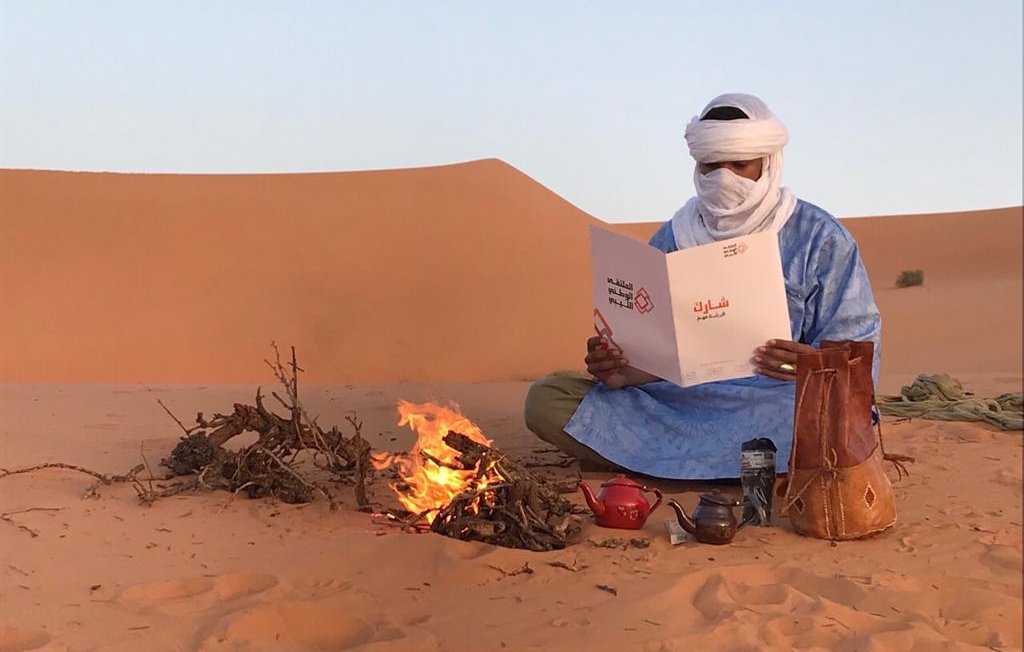 L'image montre un homme assis dans le désert, portant des vêtements traditionnels bleus et un turban. Il est devant un petit feu de camp, où des brindilles brûlent. L'homme tient un menu qu'il examine attentivement. À côté de lui, il y a une théière et un pot en terre cuite. En arrière-plan, on peut voir des dunes de sable sous un ciel clair. L'ambiance évoque la tranquillité et la culture nomade.