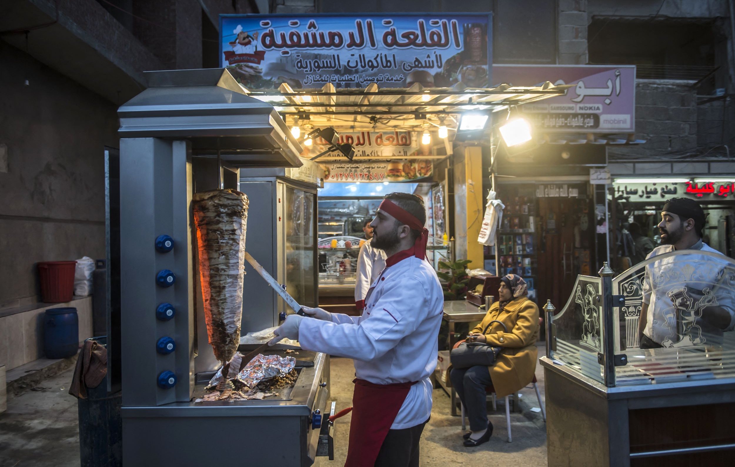 Sur cette image, on peut voir un vendeur de nourriture dans une rue animée, probablement dans un pays du Moyen-Orient. Il est en train de préparer un plat cuisiné, et on peut apercevoir un grand rouleau de viande, typique des stands de kebab ou de shawarma. Le vendeur porte une tenue de chef, composée d'une veste blanche et d'un couvre-chef. À côté de lui, une autre personne semble attendre, assise et observant. L'ambiance est chaleureuse, avec des lumières attirantes au-dessus du stand et des panneaux indiquant le nom du lieu.