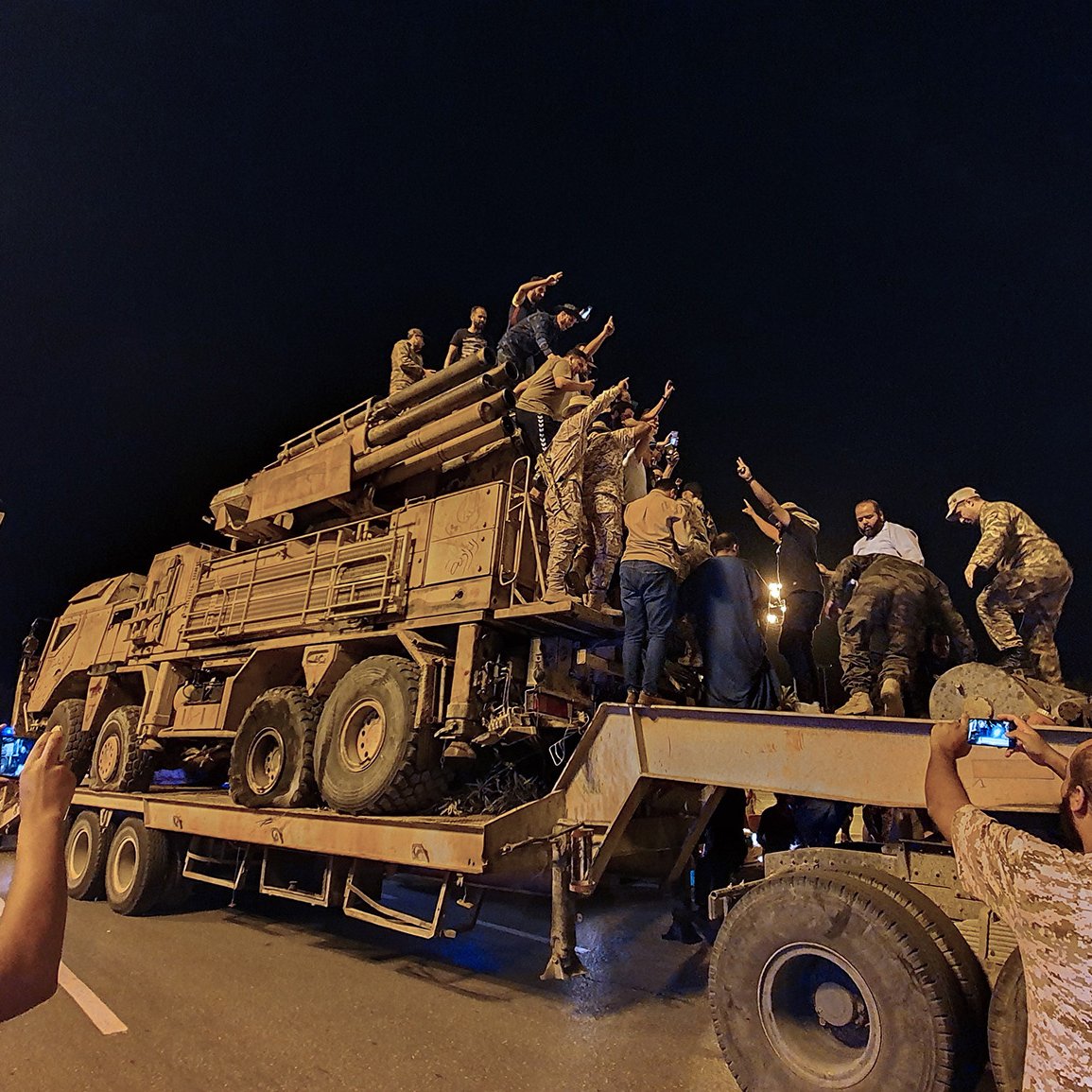 The image shows a large military vehicle, likely a truck, being used in a nighttime setting. A group of people, some in military attire and others in civilian clothing, are gathered around the vehicle. Some individuals are climbing on top of it, while others are taking photos with their phones. The atmosphere appears lively, and there is a sense of celebration or excitement among the crowd. The scene is illuminated by artificial lights, contributing to the nighttime ambiance.