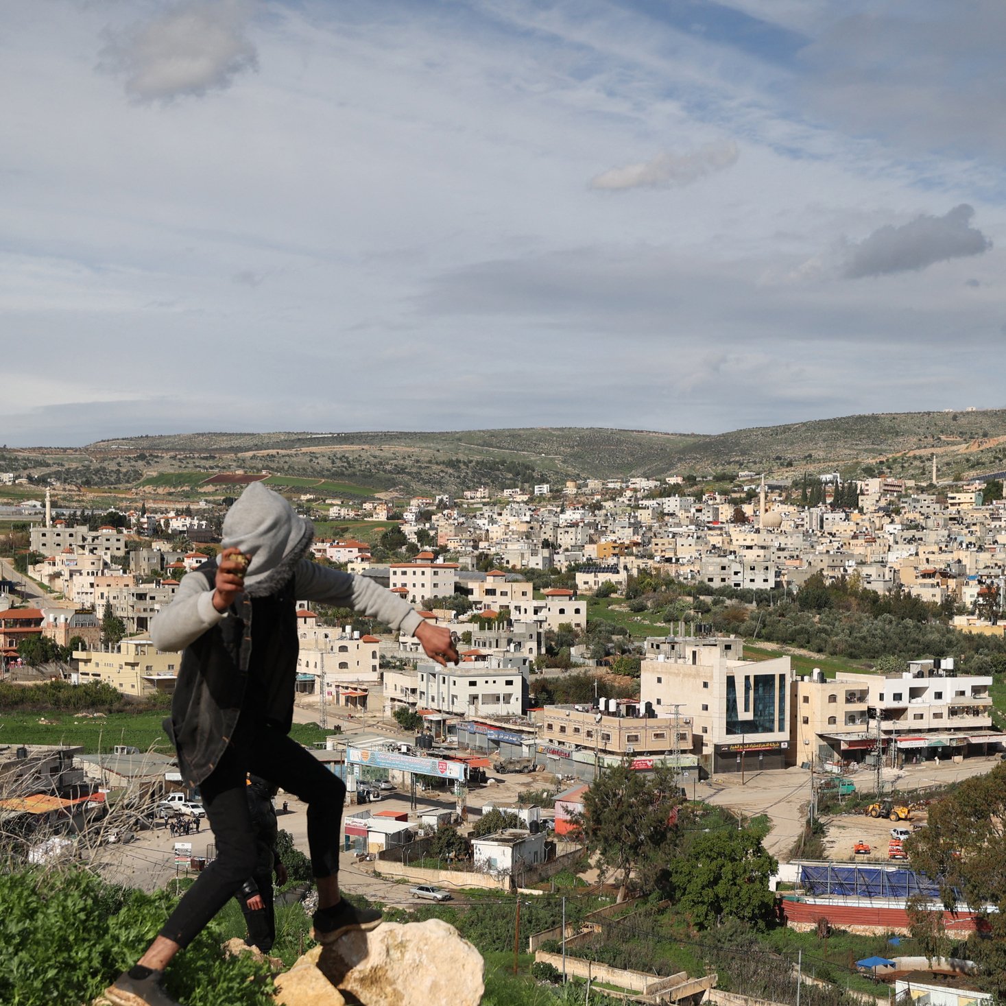 تظهر الصورة مشهداً طبيعياً لمدينة أو قرية، حيث تطل المباني السكنية على مساحات خضراء. في مقدمة الصورة، يوجد شخص يقف على صخرة، يبدو أنه في حالة حركية، بينما تظهر خلفه منظر عام للمدينة والمناطق المحيطة بها. السماء مليئة بالغيوم، مما يعكس أجواء متغيرة. تبرز في الصورة شوارع ومزارع، مما يعطي انطباعاً عن البيئة المحلية.