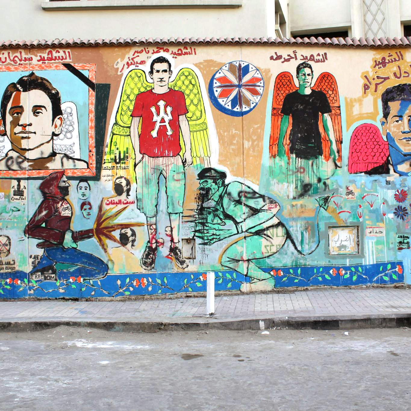 L'image représente une fresque murale colorée sur un mur. On y voit plusieurs portraits d'individus, accompagnés de motifs d'ailes et de détails symboliques. Les personnages ont des expressions variées, et des écritures en arabe sont visibles en haut de la fresque. Le fond présente des éléments artistiques tels que des fleurs et d'autres motifs graphiques. L'ambiance générale est vibrante et évoque un message fort, probablement en lien avec la culture locale ou un hommage à des figures importantes. Le sol devant le mur est en béton, et on peut y voir des supports en métal.