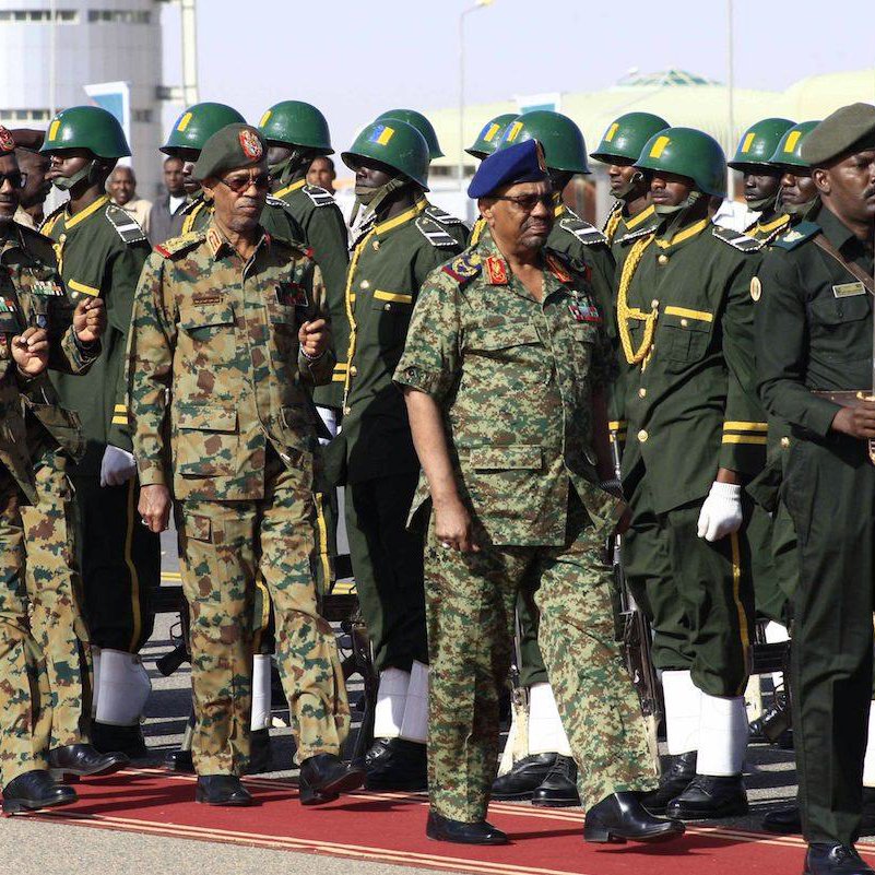 L'image montre un groupe de militaires en uniforme, marchant en formation. Les soldats portent des uniformes variés, notamment des uniformes camouflés et d'autres de couleur kaki, ornés de médailles et d'insignes. Certains militaires portent des casques ou des bérets. Au premier plan, des officiers semblent inspecter les troupes, tandis qu'une formation de soldats en arrière-plan est prête, alignée sur une surface rouge, probablement lors d'une cérémonie officielle. L'ambiance semble solennelle et rituelle, caractéristique d'une parade militaire.