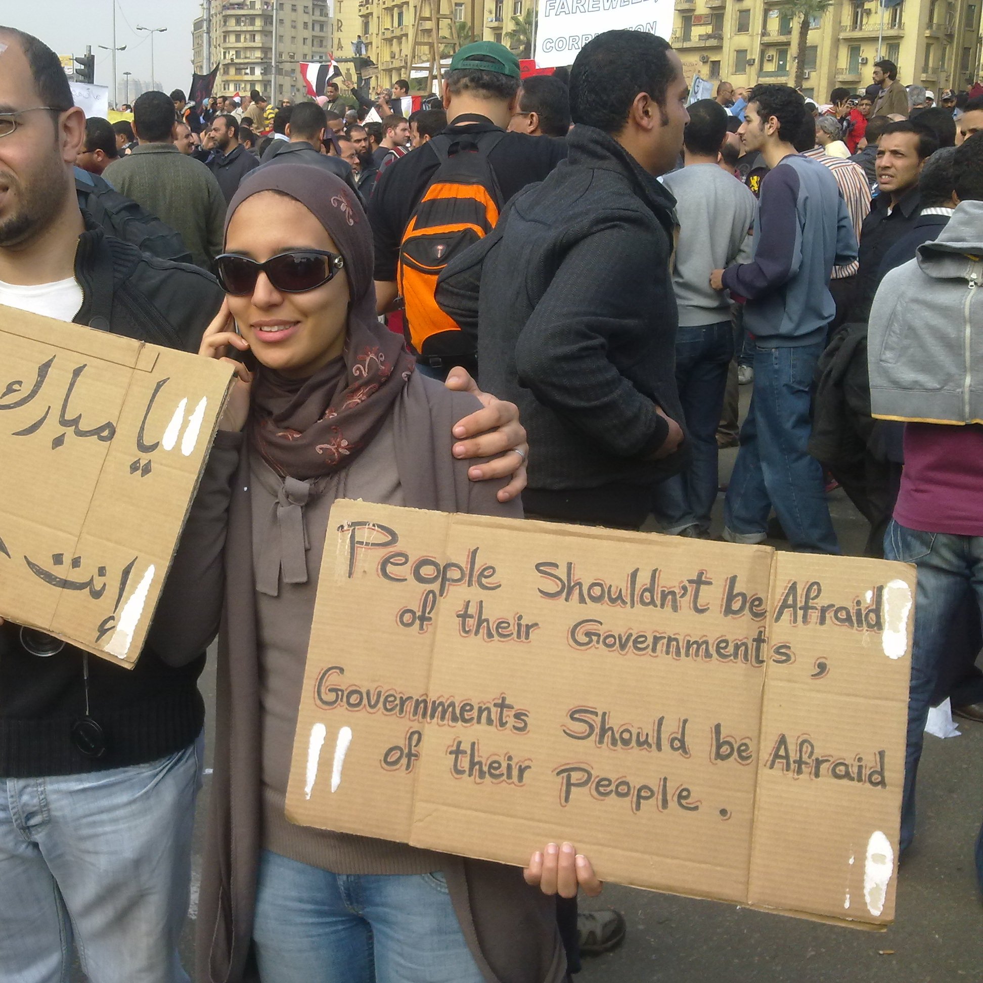 L'immagine mostra un gruppo di persone in una manifestazione. Due individui in primo piano tengono in mano dei cartelli. Uno dei cartelli è scritto in arabo e l'altro in inglese. Il messaggio in inglese dice "Le persone non dovrebbero avere paura dei loro governi. I governi dovrebbero avere paura del loro popolo." I partecipanti sembrano impegnati e determinati, con un'ampia folla di persone sullo sfondo. La scena evoca un senso di protesta e richiesta di cambiamento sociale.