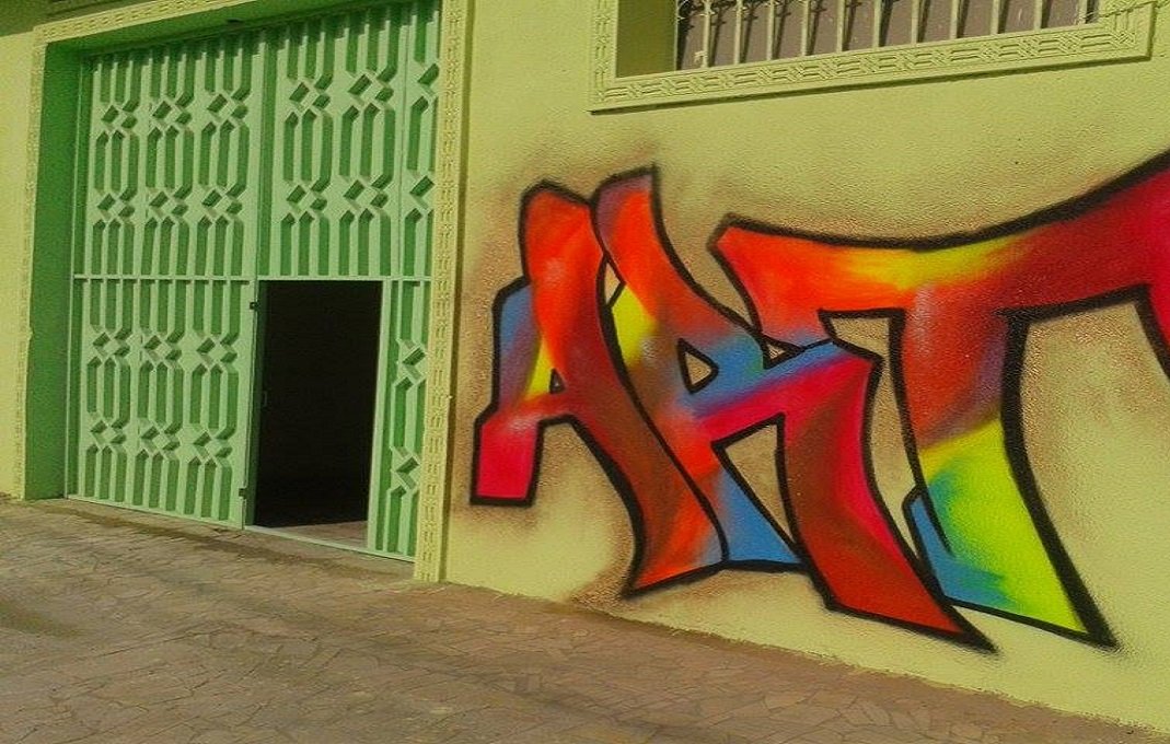 الصورة تظهر جدارًا ملونًا يحمل رسمة جرافيتي تحتوي على كلمة "ART" بألوان زاهية مثل الأحمر والأصفر والأزرق. الجدار هو بجانب باب أخضر فاتح، مما يضيف لمسة مميزة ومبهجة إلى المشهد. التصميم يعكس روح الفن والإبداع في البيئة المحيطة.