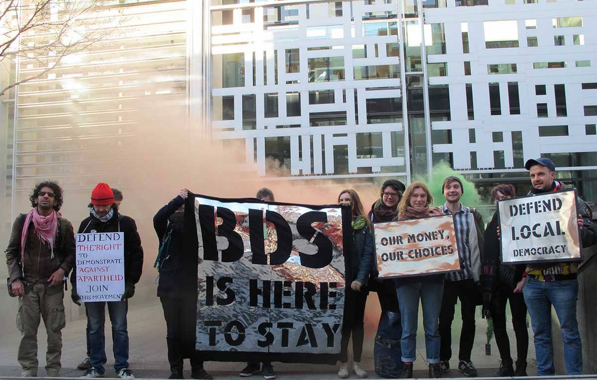 L'image montre un groupe de personnes mobilisées lors d'une manifestation. Elles tiennent différentes pancartes avec des messages engagés, notamment en soutien au mouvement BDS (Boycott, Désinvestissement, Sanctions). L'ambiance semble dynamique et militante, avec des couleurs vives et une légère fumée en arrière-plan, ce qui suggère un moment de tension ou d'énergie. Les participants, vêtus de manière variée, affichent des expressions déterminées face à la caméra.