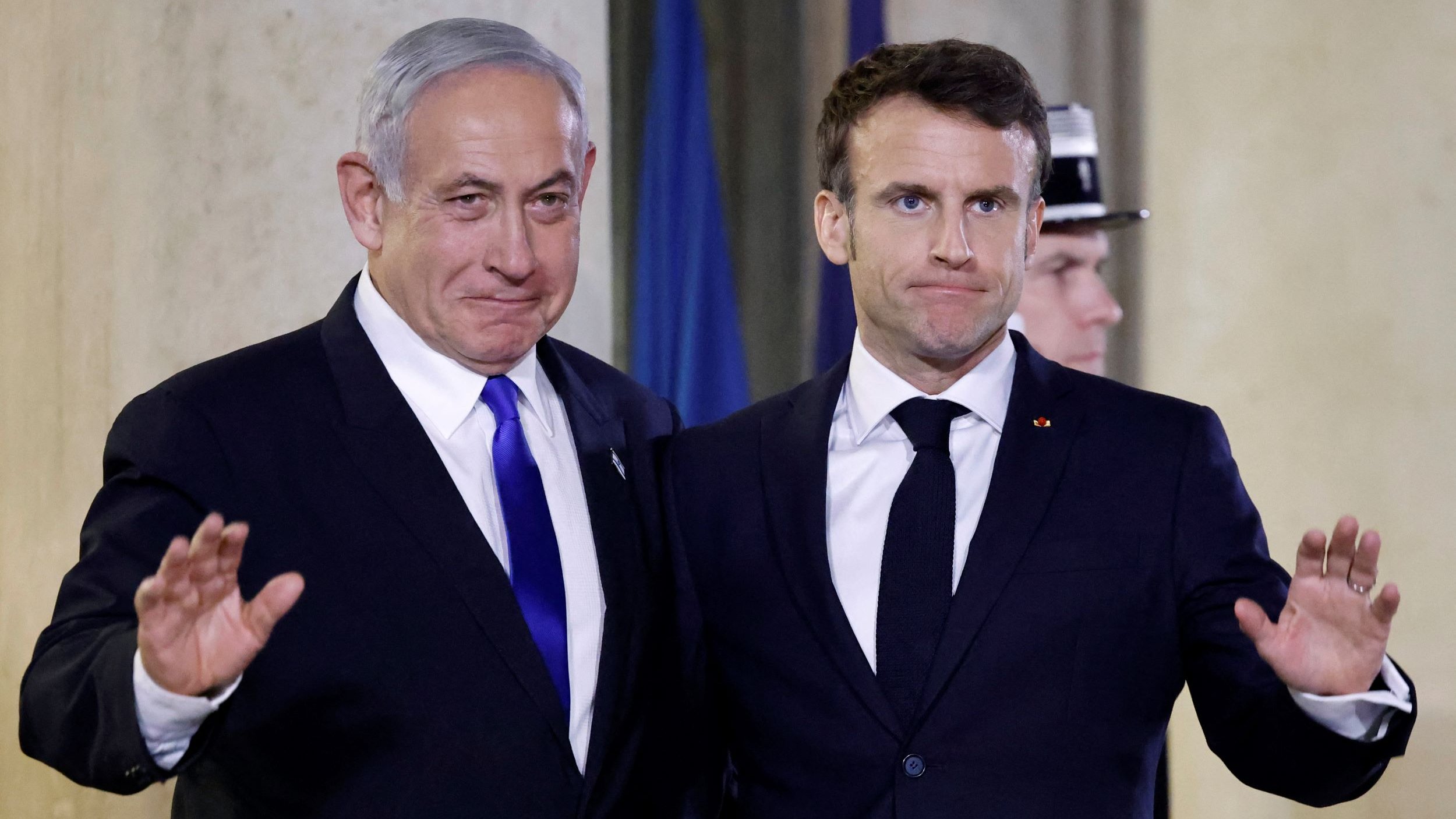 L'image montre deux hommes, l'un en costume sombre et cravate bleue, l'autre également en costume sombre mais avec une cravate noire. Ils se tiennent côte à côte, levant la main dans un geste de salutation. L'arrière-plan suggère un cadre officiel, possiblement lors d'une rencontre ou d'une conférence de presse. Les deux semblent sérieux.