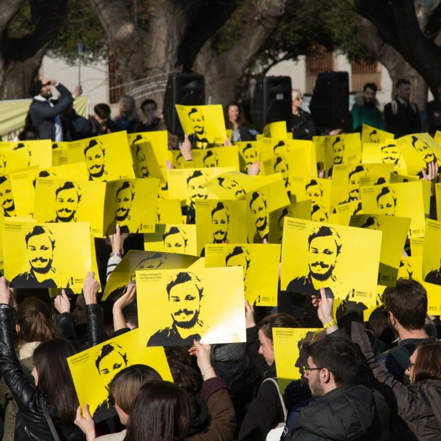 L'image montre une grande foule rassemblée, tenant des pancartes jaunes avec un visage représenté en noir. Les participants semblent mobilisés pour une cause, se tenant la main levée en signe de protestation ou de soutien. En arrière-plan, on peut voir d'autres personnes et des arbres, suggérant que l'événement a lieu en plein air. L'atmosphère paraît engagée et déterminée.