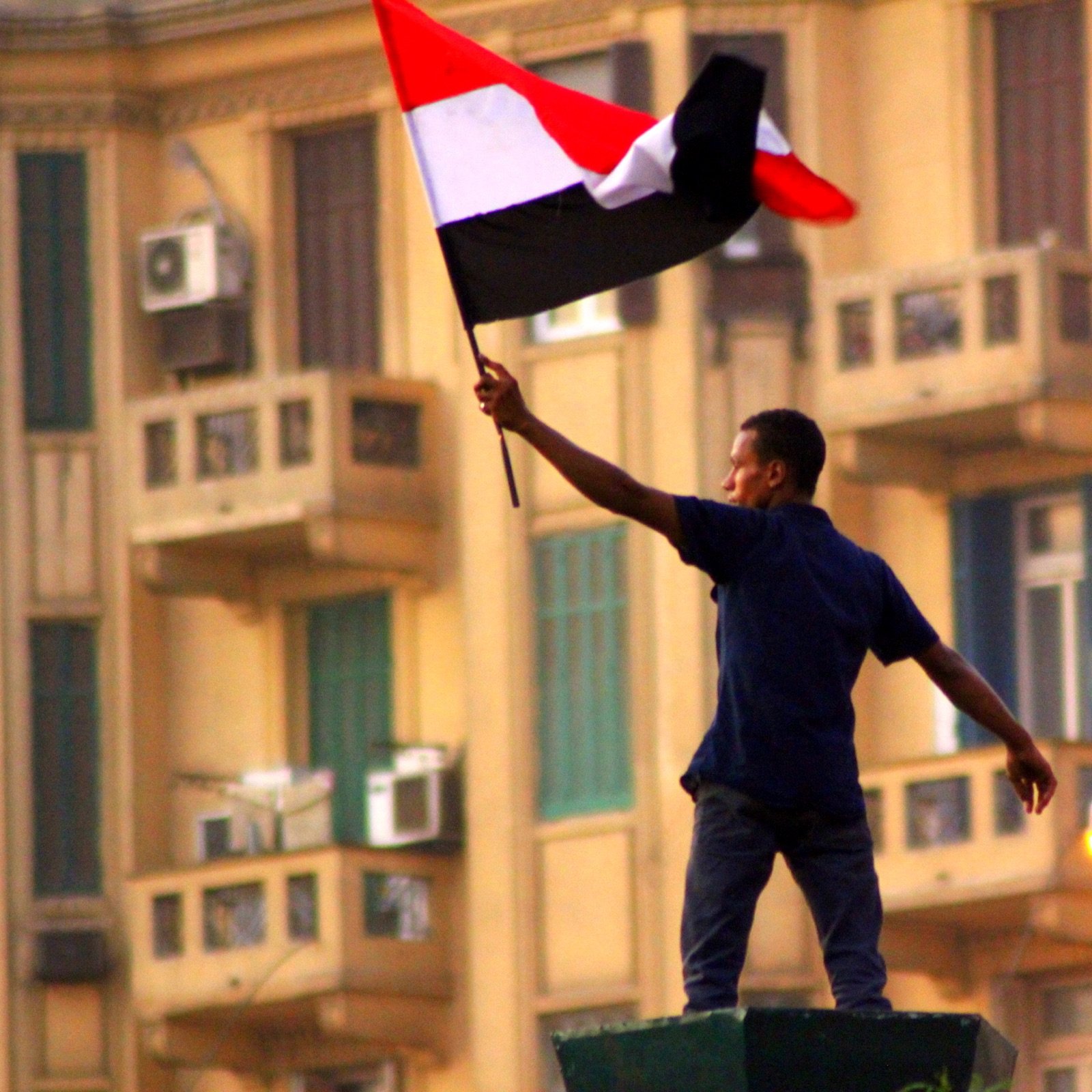 L'image montre un homme brandissant un drapeau noir, blanc et rouge, probablement un drapeau national. Il se tient debout sur une structure, manifestant une posture de célébration ou de protestation. En arrière-plan, on peut apercevoir des bâtiments avec des balcons et des climatiseurs, ce qui suggère un environnement urbain. L'atmosphère semble chargée d'une forte émotion, possiblement liée à un événement politique ou social.