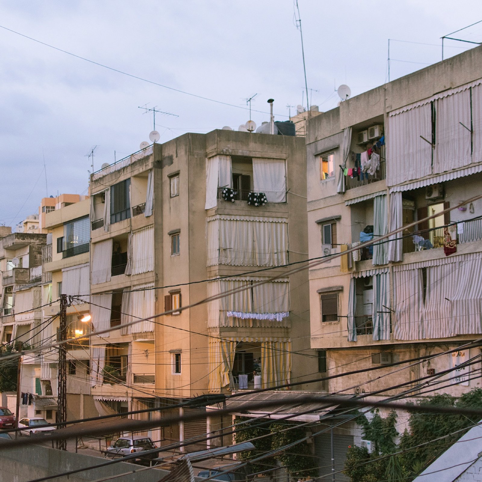 L'image montre un ensemble d'immeubles résidentiels dans une zone urbaine. Les bâtiments semblent anciens et sont de plusieurs étages, avec des balcons où l'on peut voir des vêtements étendus. La lumière du soir crée une atmosphère douce, et des fils électriques traversent l'image. On aperçoit également des voitures garées le long de la rue en contrebas. L'ensemble donne une impression de vie citadine typique, avec un mélange de modernité et de caractère ancien.