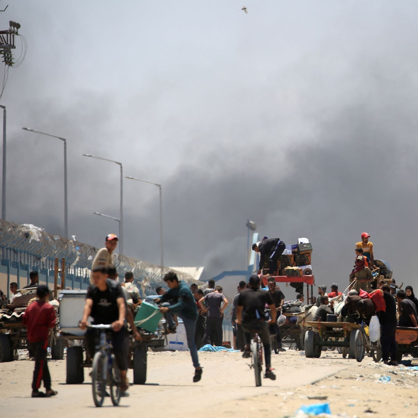 تصویر یک صحنه شهری را نشان می‌دهد که در آن تعدادی از افراد در حال حرکت هستند. در زمینه، دود سیاهی به آسمان بلند شده که نشان‌دهنده وجود یک حادثه یا آتش‌سوزی است. افرادی که در تصویر دیده می‌شوند، شامل مردان، زنان و کودکانی هستند که برخی از آنها در حال دوچرخه‌سواری و برخی دیگر در حال حمل بار در کالسکه‌های دستی هستند. فضای کلی تصویر به نظر شلوغ و پر از فعالیت است.