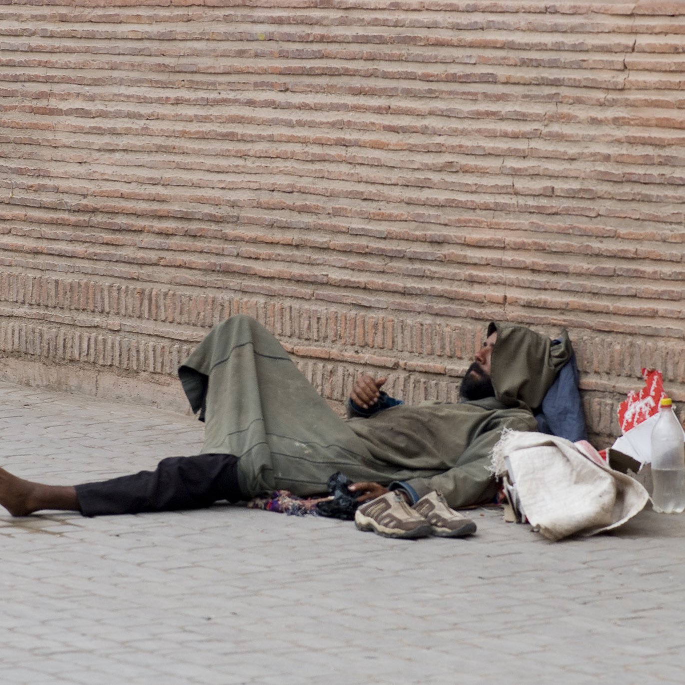 L'image montre une personne allongée sur le sol, semblant sans abri. Elle porte des vêtements sombres et usés, et se trouve à côté d'une paroi de briques. On voit des objets éparpillés autour d'elle, comme des chaussures et un sac. L'environnement semble urbain et le ton de l'image est contemplatif.