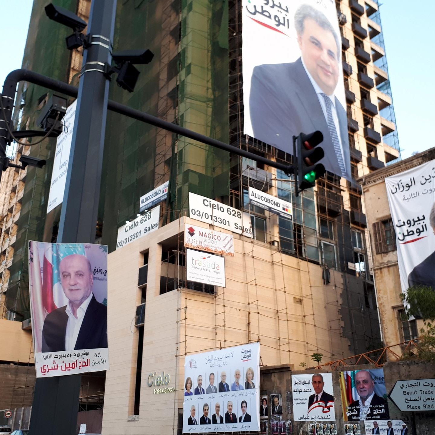 L'image montre un immeuble en construction ou rénové, arborant plusieurs affiches et panneaux publicitaires. Les affiches présentent des portraits de différentes personnes, probablement des candidats ou des figures politiques, accompagnés de textes en arabe. La scène donne l'impression d'une campagne électorale ou d'une promotion politique. Il y a également un feu de signalisation visible à gauche, ce qui indique que cette image est prise dans un environnement urbain.