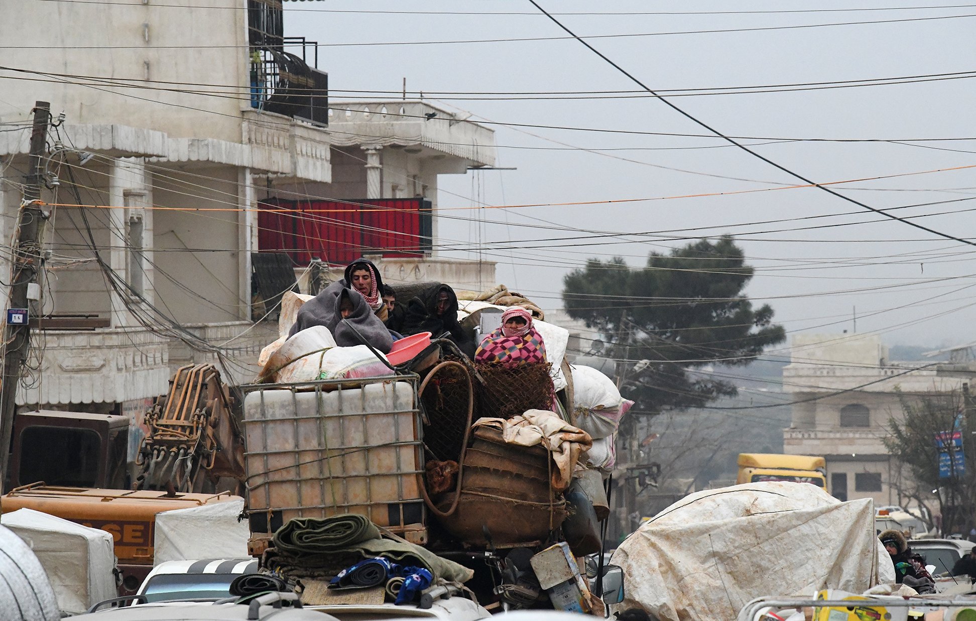 L'image montre une scène urbaine chargée, où des personnes sont perchées sur une pile de marchandises. Elles semblent se déplacer sur un véhicule surchargé, avec différents types de bagages et de matériaux. L'environnement est voilé par une lumière tamisée, peut-être due à un temps nuageux ou brumeux, et des câbles électriques traversent la scène. Les bâtiments en arrière-plan suggèrent un contexte urbain. Cette image évoque un sentiment de mouvement et de survie dans un espace urbain encombré.