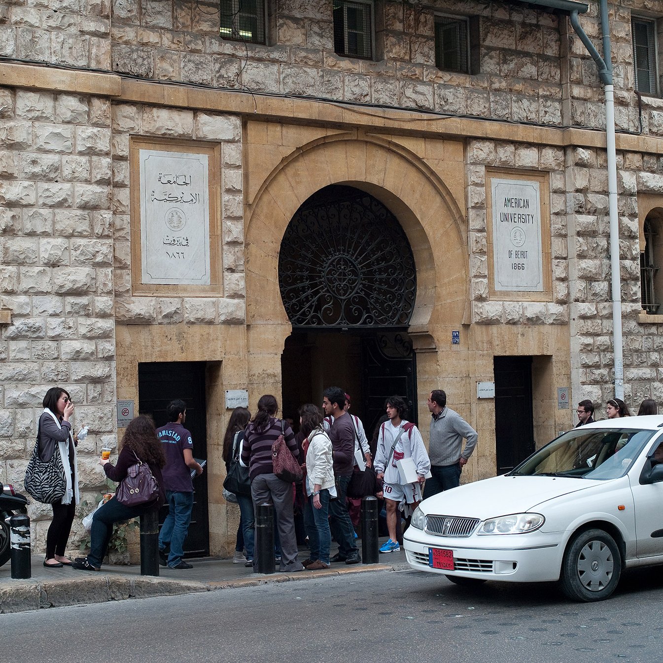 L'image montre l'entrée d'un bâtiment en pierre, probablement une université. Plusieurs personnes sont rassemblées devant l'entrée, créant une petite file d'attente. On peut voir des étudiants, certains portant des sacs. À droite, une voiture blanche passe proche de la scène, tandis qu'une moto est garée à proximité. L'architecture du bâtiment présente des éléments classiques avec un grand portail en fer.
