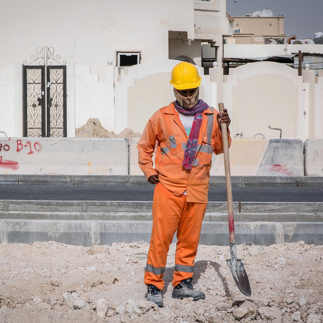 Nell'immagine si vede un lavoratore in un cantiere edile. Indossa una tuta arancione e un casco giallo per la sicurezza. Ha in mano una pala e sembra essere in pausa, osservando il terreno di fronte a lui. Sullo sfondo si possono notare alcune strutture e graffiti su un muro. L'ambiente appare polveroso e in fase di costruzione.