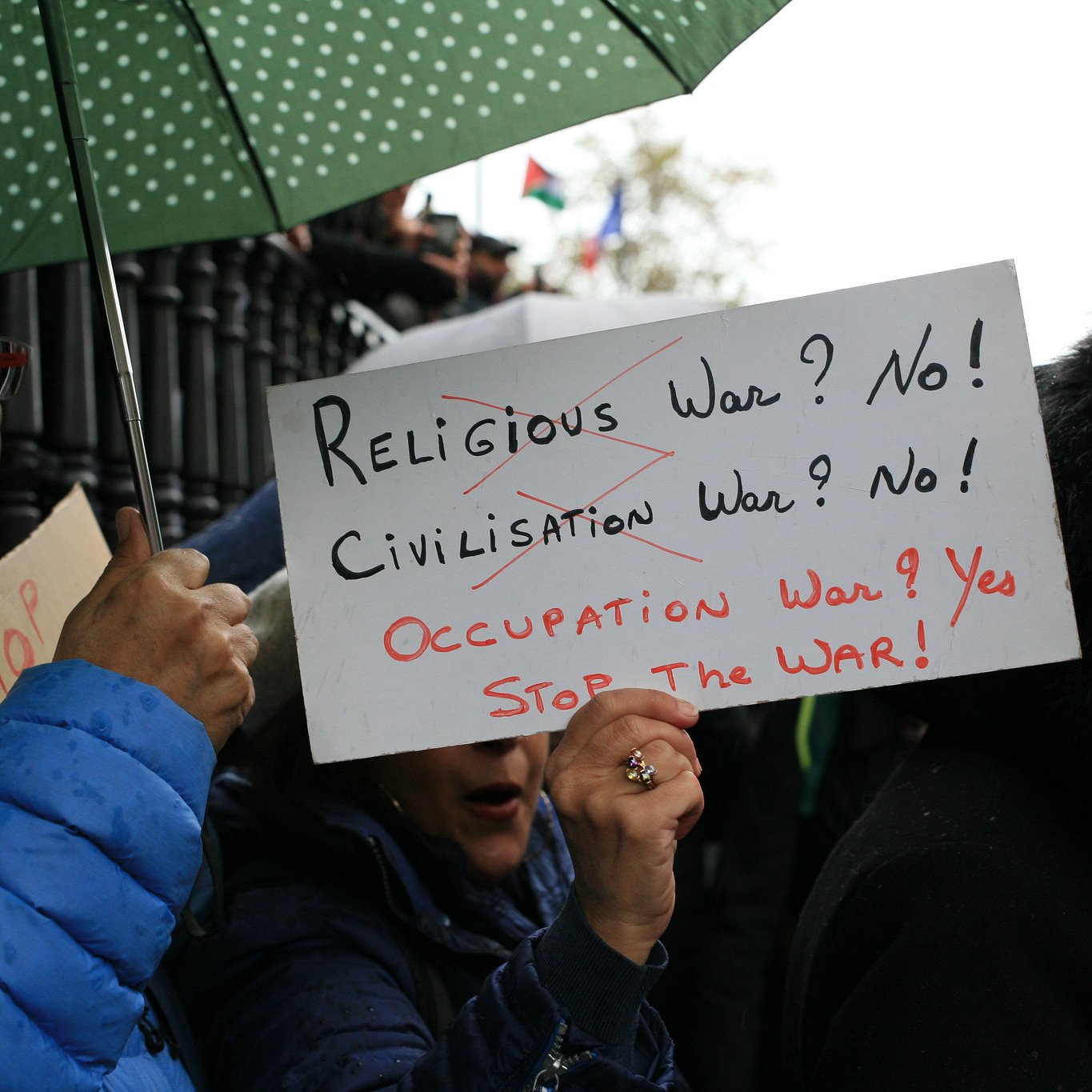 في الصورة، يظهر مجموعة من الأشخاص في تظاهرة، حيث يحمل أحدهم لافتة مكتوب عليها "الحرب الدينية؟ لا! الحرب الحضارية؟ لا! حرب الاحتلال؟ نعم! اوقفوا الحرب!"، بينما يحمل آخر مظلة خضراء. التعبيرات على وجوه المتظاهرين تدل على الإصرار والمشاركة الفعالة في الاحتجاج. الأجواء تتسم بالجدية مع خلفية غائمة أو ممطرة.