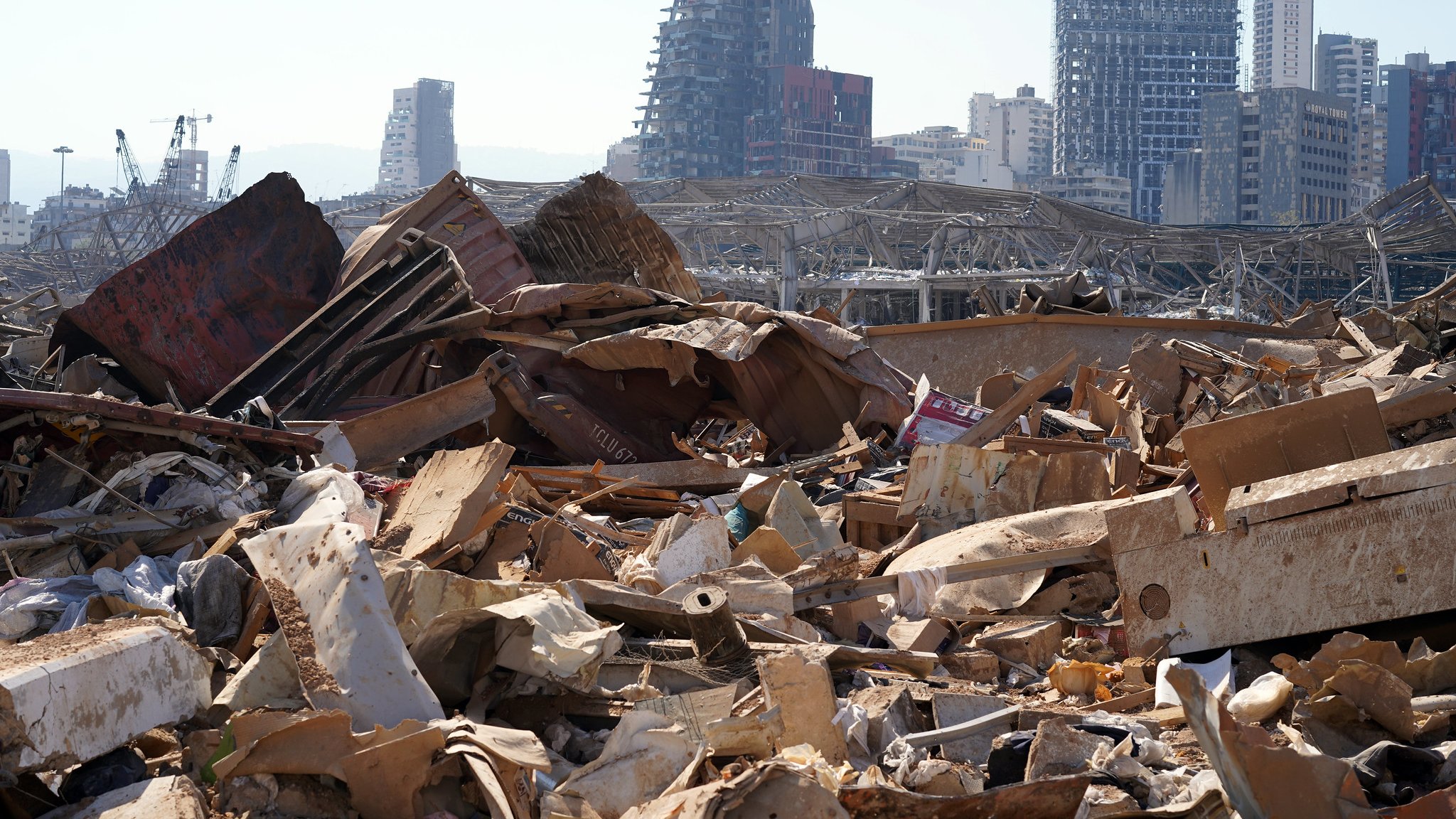 L'image montre un paysage urbain dévasté, avec des débris et des matériaux en désordre dans le premier plan. On peut voir des bâtiments partiellement détruits en arrière-plan, témoignant des dommages importants. L'atmosphère semble désolée, illustrant l'impact d'une catastrophe, probablement une explosion ou une destruction massive. Des structures modernes sont visibles, mais leur état montre les effets néfastes de l'événement. Le ciel est dégagé, contrastant avec la gravité de la scène au sol.