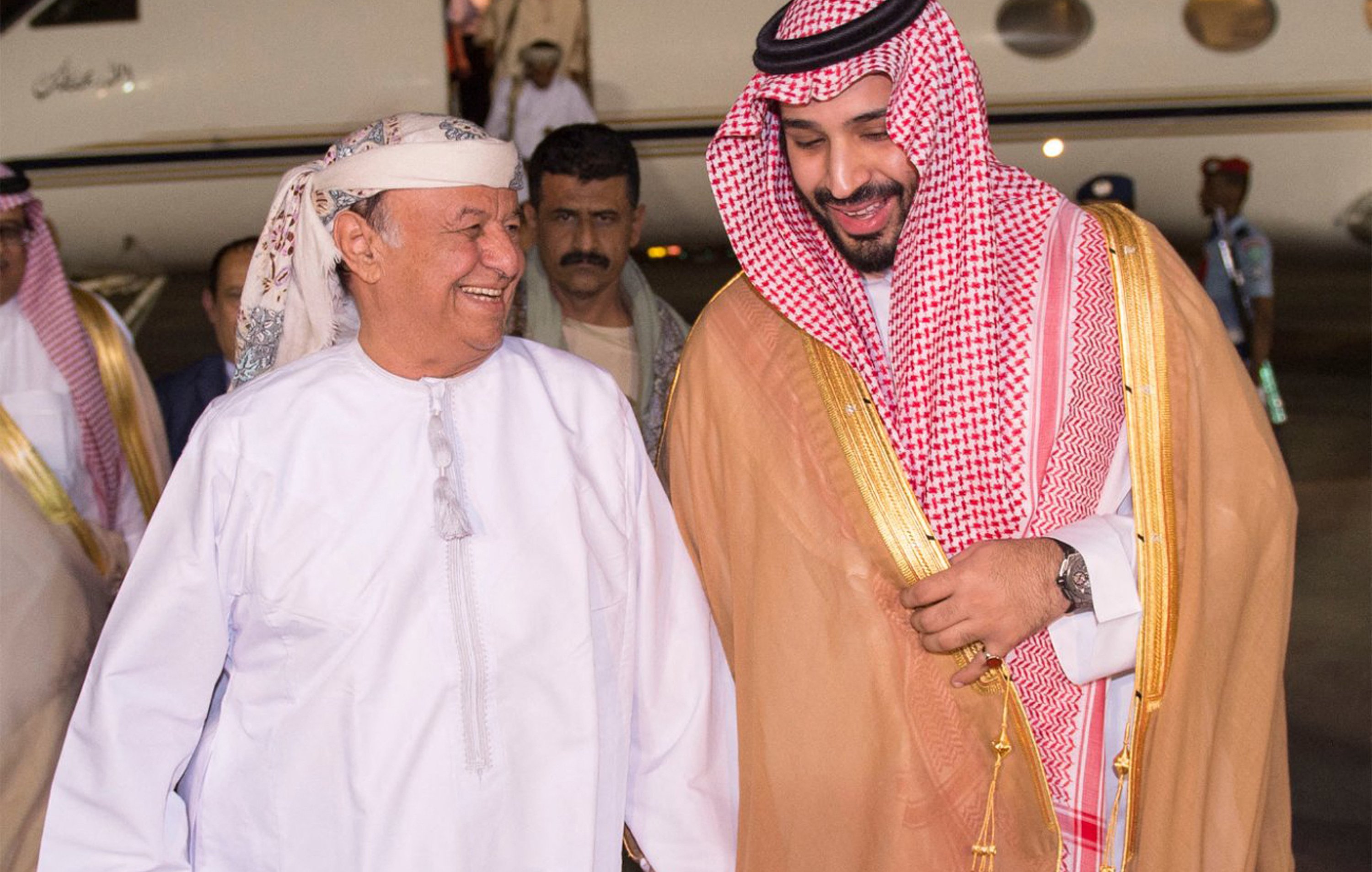 The image shows two men smiling and engaged in a friendly conversation as they walk towards a private jet. One man is dressed in traditional Arab attire, including a white thobe and a headscarf, while the other is wearing a similar traditional outfit, accented with a golden cloak and a checkered headscarf. They appear to be happy and are holding hands, suggesting a warm greeting. In the background, there are several individuals, likely staff or security, standing by the jet. The setting seems to be an airport or a private aviation terminal.