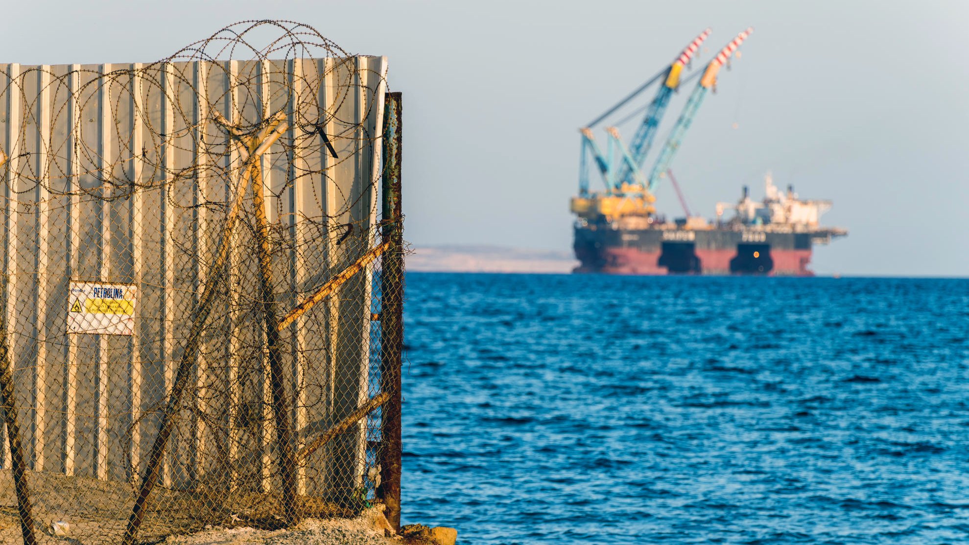 L'image montre une barrière en métal, probablement un mur de séparation, avec du fil barbelé à sa base. En arrière-plan, on aperçoit des barges ou des plateformes offshore, avec des grues, sur un fond d'eau calme. Le ciel semble dégagé, et l'ensemble donne une impression de contraste entre la structure humaine et l'environnement maritime.