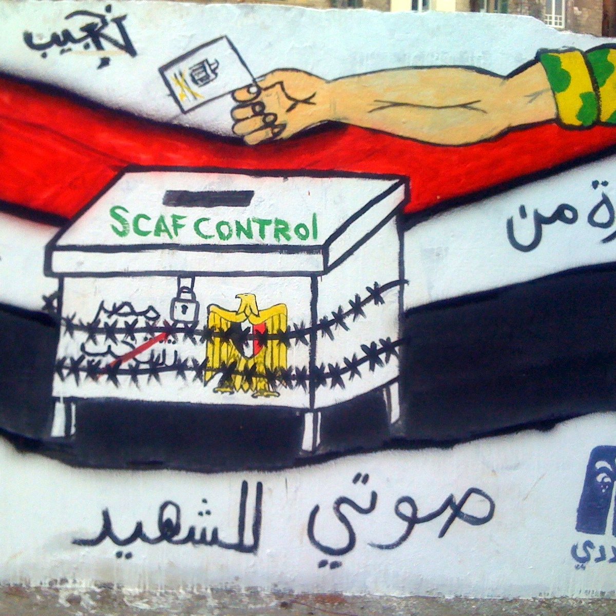 L'image montre une fresque murale avec une boîte de vote entourée de barbelés. Au-dessus de la boîte, on peut voir la phrase "SCAF CONTROL" écrite en vert. À gauche, il y a un poing levé, symbolisant la résistance ou la lutte, et à droite, on peut apercevoir des symboles qui représentent probablement des idéaux de liberté et de changement. La fresque utilise des couleurs vives comme le noir, le rouge et le blanc, qui sont souvent associés à des mouvements politiques. Le message semble critiquer ou commenter le contrôle militaire sur le processus électoral.