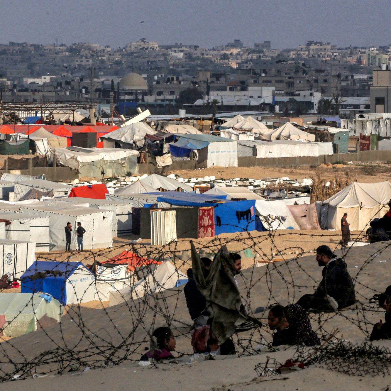 این تصویر منطقه‌ای را نشان می‌دهد که پر از چادرهای سفید و آبی است. در پس‌زمینه، ساختمان‌های شهری دیده می‌شود. جمعی از افراد در نزدیکی چادرها نشسته‌اند و به نظر می‌رسد که در یک وضعیت دشوار زندگی می‌کنند. همچنین، نوارهایی از سیم‌خاردار در عکس به چشم می‌خورد که نشان‌دهنده محدودیت‌های امنیتی است. فضای کلی تصویر حاکی از چالش‌های انسانی و وضعیت اضطراری است.