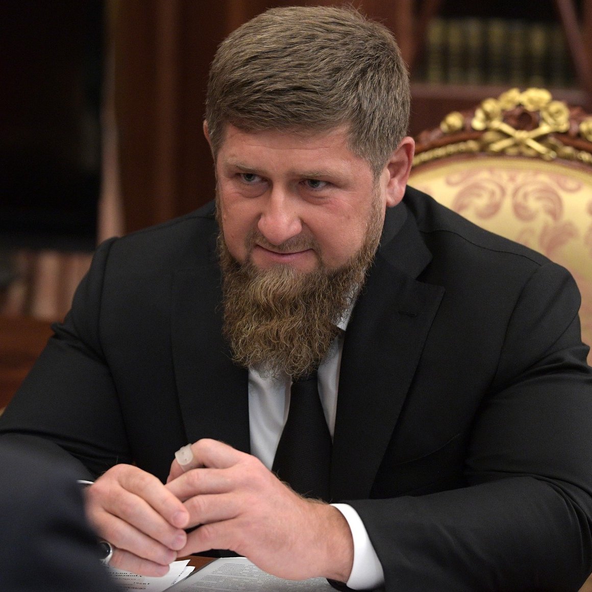 L'image montre un homme avec une barbe, assis à une table dans un cadre formel. Il est habillé en costume noir et semble concentré sur la discussion. En arrière-plan, on peut apercevoir des meubles élégants et une ambiance sophistiquée. L'éclairage est doux, ce qui contribue à l'atmosphère sérieuse de la scène.