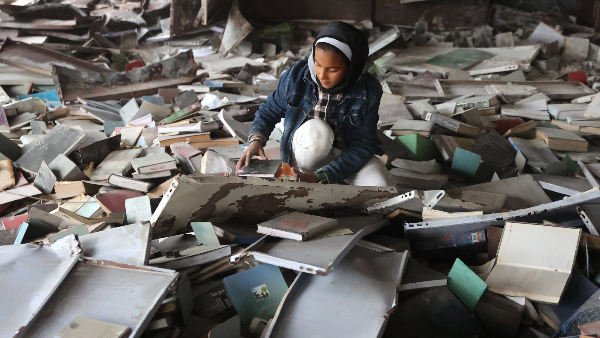 L'image montre une jeune fille assise au milieu d'un tas de livres et de débris. Elle semble explorer et manipuler les objets autour d'elle dans un environnement en désordre.