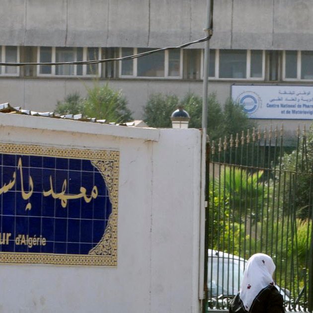 L'image montre l'extérieur de l'Institut Pasteur en Algérie. On peut voir un bâtiment moderne en arrière-plan avec des panneaux d'affichage. À l'avant-plan, il y a une entrée avec une façade blanche où figure le nom de l'institut en caractères arabes et français. Quelques personnes marchent dans cette zone, suggérant un environnement fréquenté. Des arbres et de la végétation ajoutent une touche de verdure au décor.