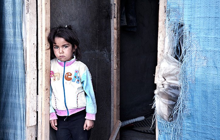 L'image montre une petite fille se tenant près d'une porte dans un environnement très modeste. Elle a les cheveux longs et bruns, et porte une veste aux couleurs vives avec des décorations. Son expression semble sérieuse et pensive. La porte et les murs environnants sont faits de matériaux simples, suggérant une situation de vie difficile. Cela évoque des thèmes de vulnérabilité et de résilience.