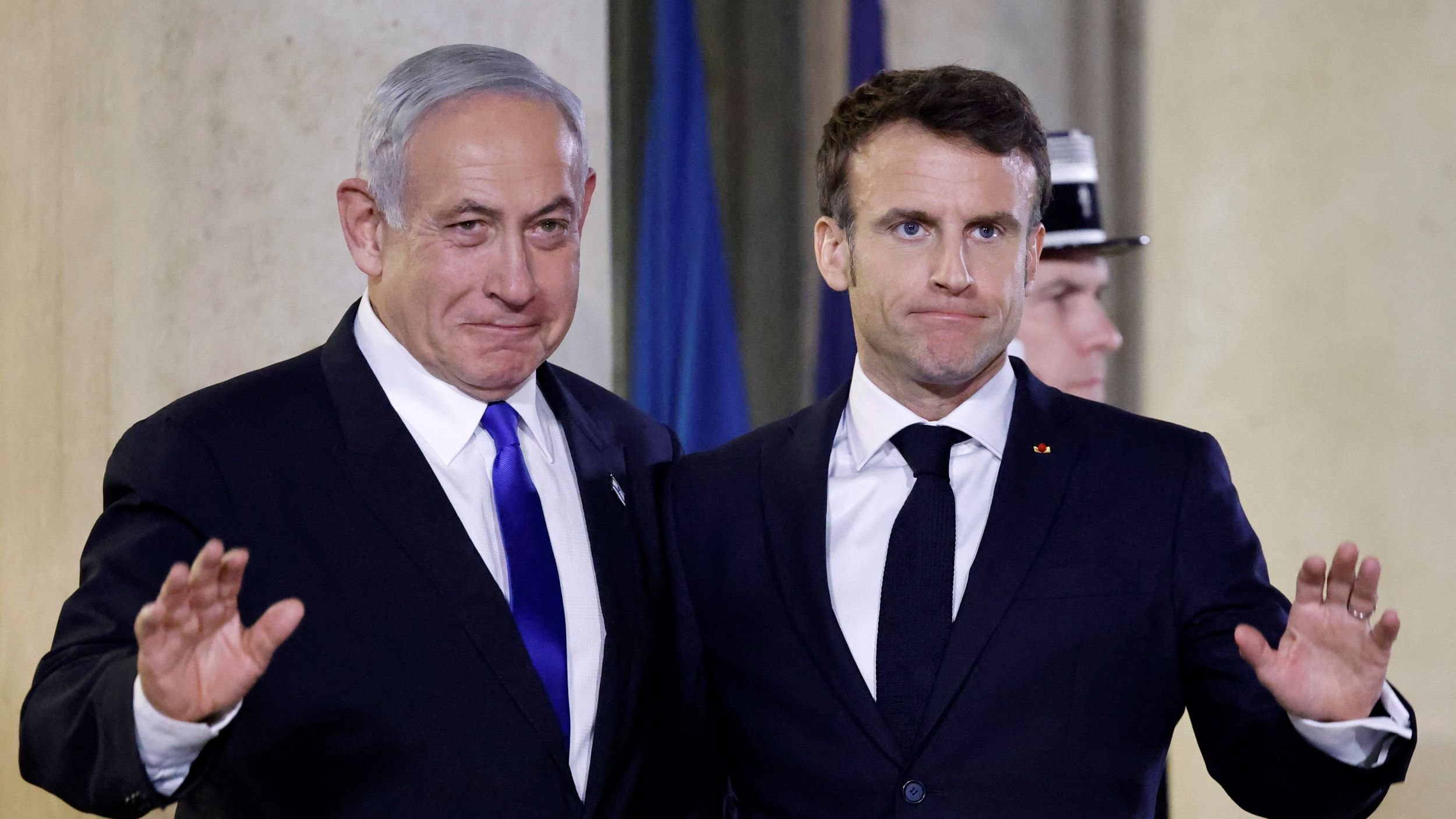 در این تصویر، دو نفر در یک موقعیت رسمی و با لبخند در کنار یکدیگر ایستاده‌اند. یکی از آن‌ها کت و کراوات تیره‌ای به تن دارد و دیگری نیز لباسی مشابه بر تن دارد. هر دو نفر به سمت دوربین نگاه می‌کنند و دستان خود را به حالت خوش‌آمد گویی بالا برده‌اند. پس‌زمینه تصویر ممکن است شامل نمادها یا پرچم‌های کشورها باشد. این تصویر به نظر می‌رسد که در یک رویداد رسمی یا کنفرانس گرفته شده است.