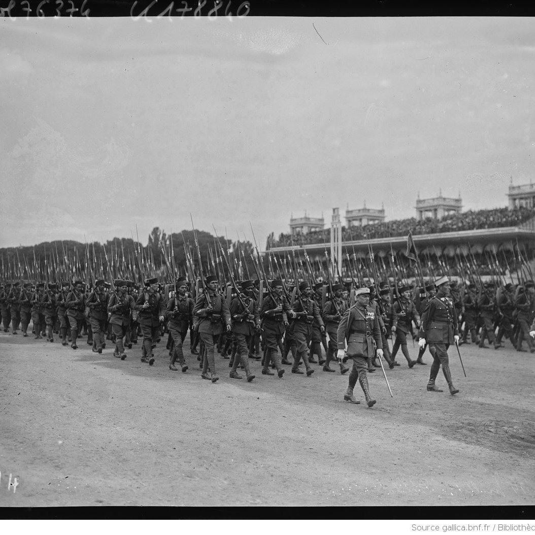 L'image montre un défilé militaire, avec de nombreux soldats marchant en formation. Ils portent des uniformes d'époque et tiennent des fusils à la main. En arrière-plan, on peut voir une tribune avec des spectateurs, probablement des dignitaires ou des spectateurs admirant le défilé. L'atmosphère semble solennelle et organisée, typique des événements militaires de ce genre. Le ciel est nuageux, ce qui donne une ambiance sobre à la scène.