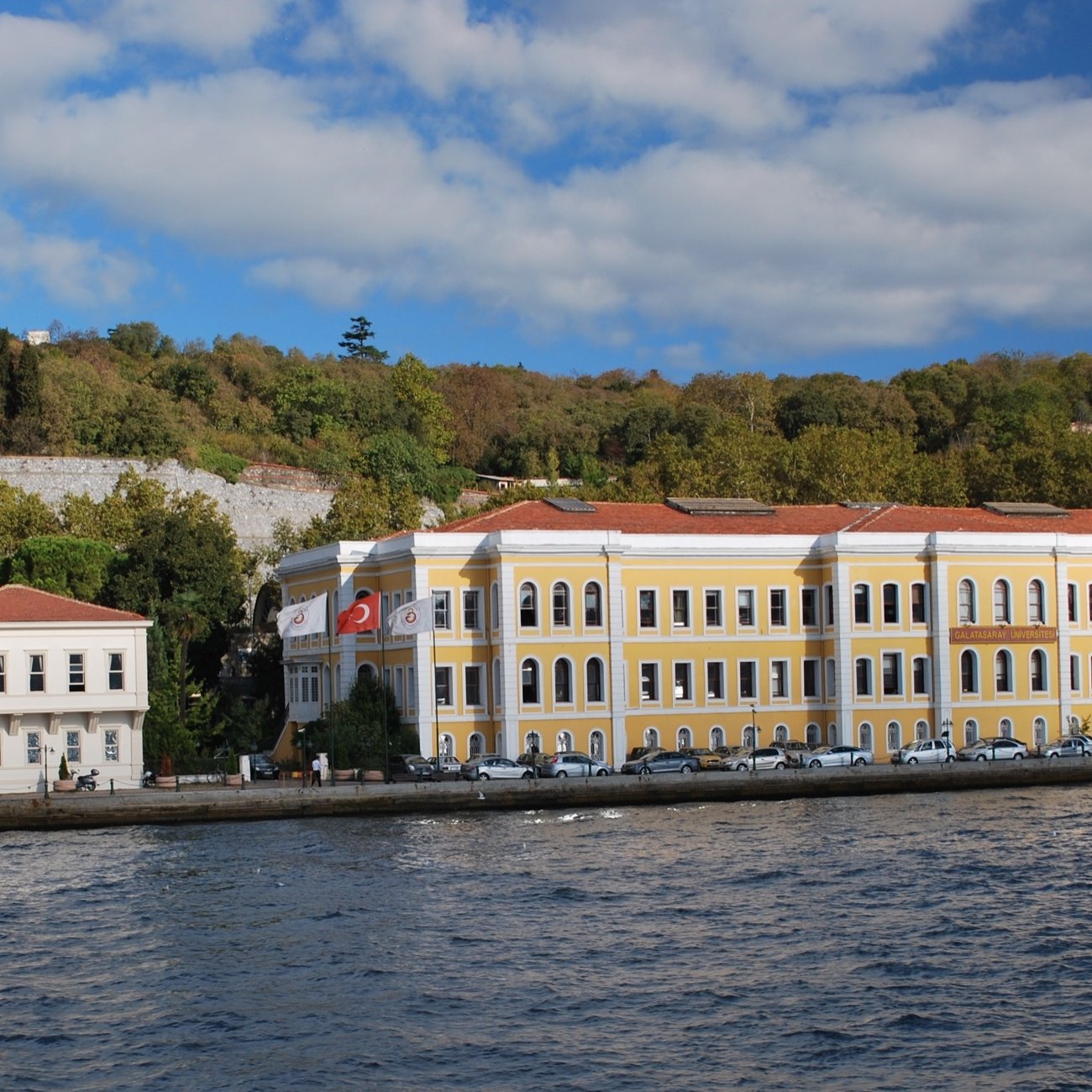 L'image montre un bâtiment jaune surplombant une étendue d'eau, probablement un bras de mer. À gauche, un grand drapeau turc flotte fièrement. On aperçoit également des arbres verdoyants en arrière-plan, ainsi qu'un ciel partiellement nuageux. Le bâtiment semble être de style architectural ancien, avec des fenêtres à volets et un toit en tuiles. Il y a des voitures garées le long de la rue en face du bâtiment. L'ensemble dégage une atmosphère paisible et pittoresque.