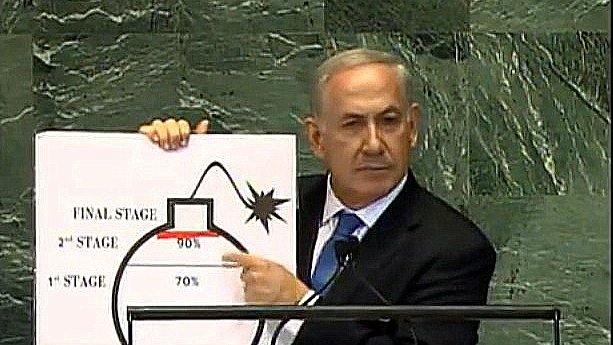 L'image montre un homme en costume, debout devant un pupitre, tenant une grande affiche. Il pointe vers un diagramme qui illustre les différentes étapes d'un processus, avec des pourcentages indiquant des niveaux d'avancement. Le diagramme semble représenter une bombe, avec des mentions pour les étapes "1ère", "2ème" et "étape finale". L'arrière-plan est celui d'un bâtiment de l'ONU, suggérant que cette présentation a lieu lors d'une réunion officielle.