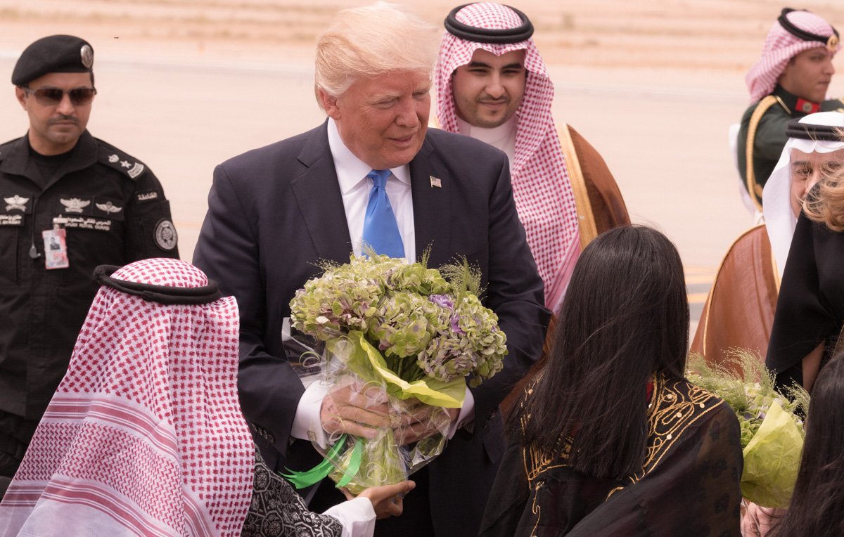 L'image montre un homme souriant, probablement un dirigeant, qui reçoit des fleurs à son arrivée. Il est entouré de plusieurs personnes portant des vêtements traditionnels, dont certains sont en tenue arabique. L'environnement semble être un aéroport, avec un paysage désertique en arrière-plan. Les personnes autour de lui semblent être accueillantes et amicales.