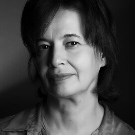 Cette image en noir et blanc montre le portrait d'une femme. Elle a les cheveux bruns et légèrement ondulés, et elle porte une chemise claire. Son expression est douce et réfléchie, et elle regarde directement l'objectif de l'appareil photo. L'éclairage met en valeur ses traits, créant un contraste entre les ombres et les lumières. L'arrière-plan est flou, ce qui accentue le visage de la femme.