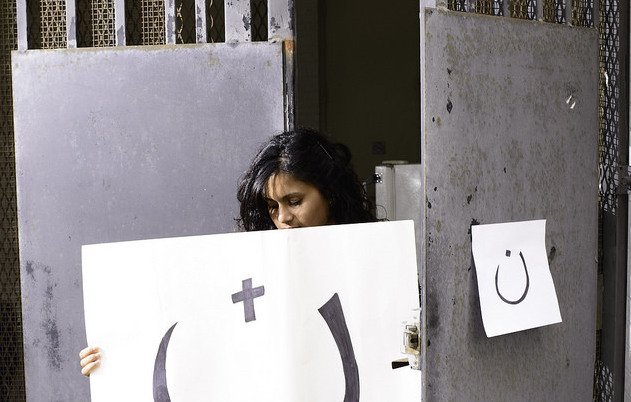 L'image montre une femme se tenant devant une porte grise. Elle tient une grande affiche présentant un symbole souriant, accompagnée d'un plus et d'un autre symbole. À côté, il y a une petite affiche accrochée à la porte. L'ensemble de la scène évoque une forme d'expression artistique, mélangeant des éléments de surprise et de réflexion.
