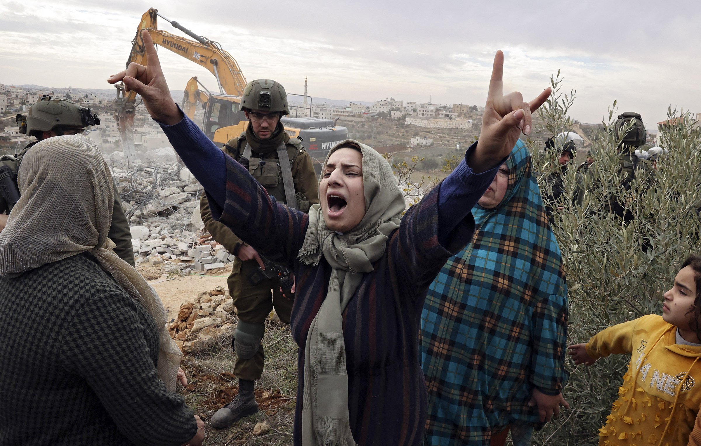 L'immagine mostra un gruppo di persone in una situazione di grande tensione emotiva. Una donna al centro, con un velo, alza le braccia in segno di protesta o di disperazione. Il suo volto esprime forte emozione. Intorno a lei, ci sono altre donne e un bambino, tutte visibilmente colpite dalla situazione. Sullo sfondo, si intravede un escavatore e una presenza militare, suggerendo che si sta svolgendo un'operazione di demolizione o sgombero. Il contesto sembra essere caratterizzato da conflitto e tensioni sociali.