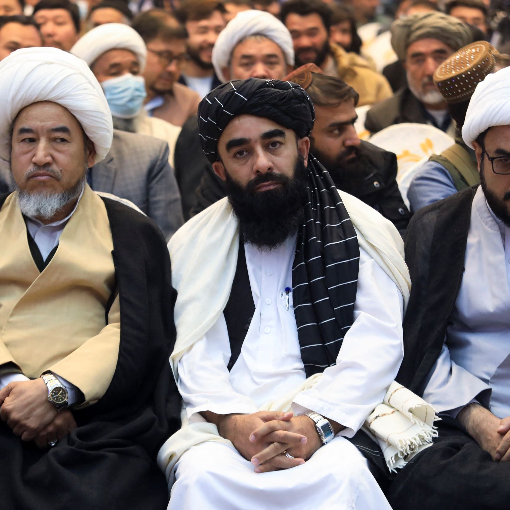 L'image montre un groupe de personnes assises, portant des vêtements traditionnels. Ils semblent participer à un événement ou une réunion. Certains d'entre eux portent des turbans ou des couvre-chefs, et l'ambiance semble solennelle. Les visages expriment diverses émotions, allant d'une concentration sérieuse à une attitude réfléchie. L'arrière-plan indique qu'il y a d'autres personnes présentes dans l'événement.