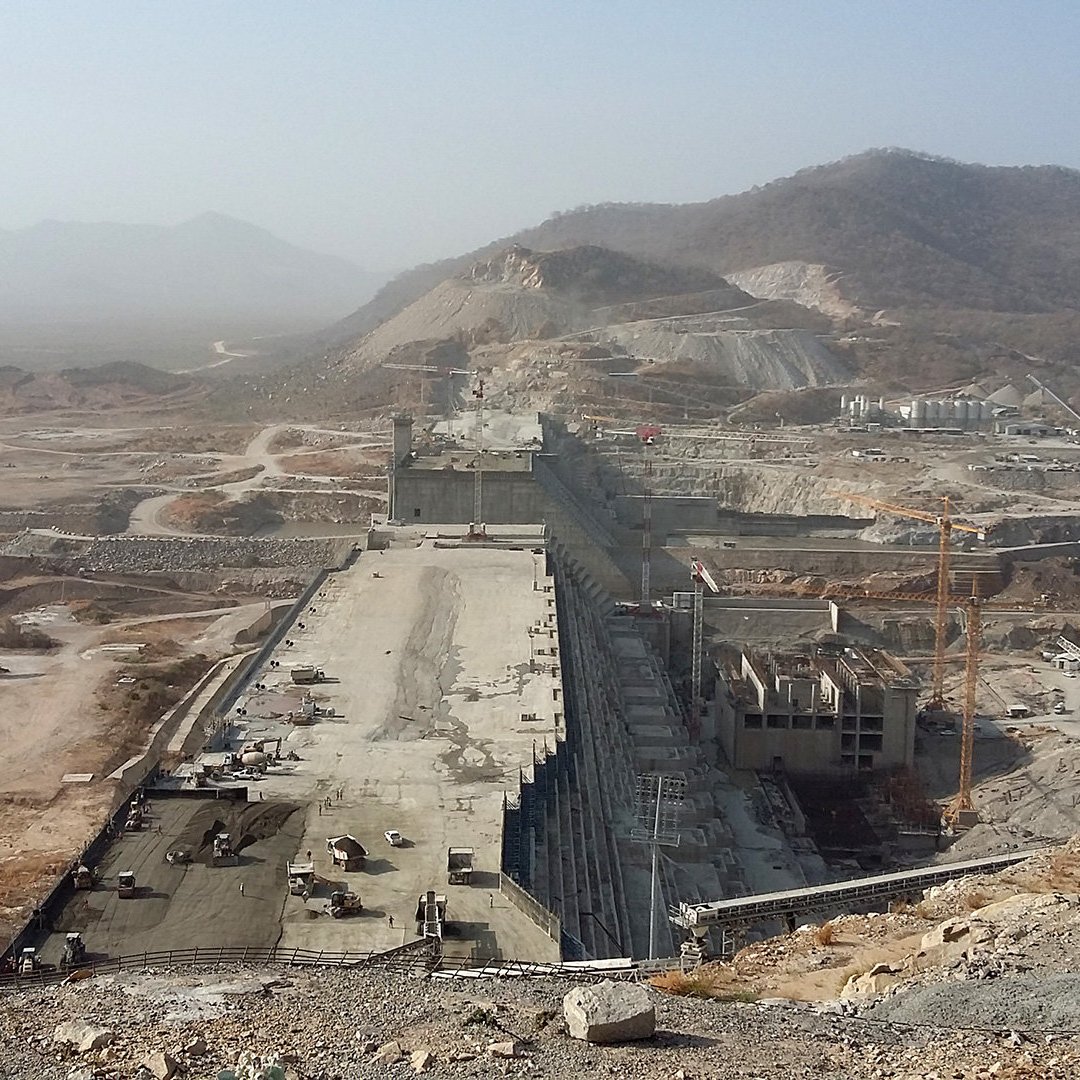 L'image montre un grand chantier de construction, probablement d'un barrage ou d'une infrastructure hydraulique. On peut voir de vastes terrassements, des routes en cours de construction et des engins lourds. L'environnement est aride, avec des collines et des montagnes à l'arrière-plan. Le ciel est légèrement brumeux, ce qui donne une atmosphère poussiéreuse à la scène. Généralement, l'image représente l'ampleur d'un projet d'ingénierie majeur.