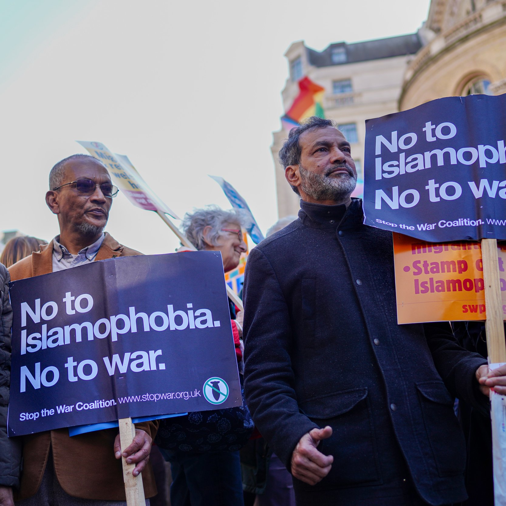 L'immagine mostra un gruppo di persone che partecipano a una manifestazione. Alcuni di loro stanno tenendo cartelli che riportano messaggi come "No to Islamophobia. No to war." e "Stop the War Coalition." L'atmosfera è di protesta pacifica, con i manifestanti che esprimono i loro punti di vista contro l'islamofobia e le guerre. In lontananza, si possono notare delle bandiere, che aggiungono un senso di diversità e unità all'evento.