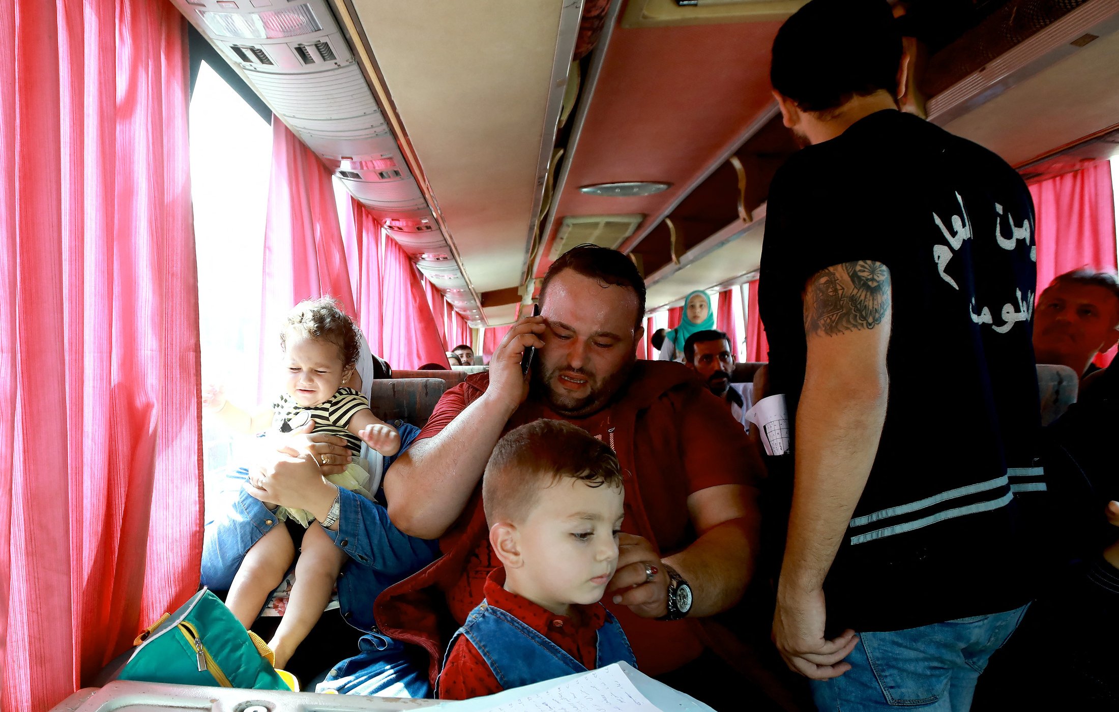 L'image montre un intérieur de bus avec plusieurs personnes. Un homme assis au centre tient un enfant dans ses bras et parle au téléphone. À ses pieds, un autre enfant semble jouer ou être attentif. D'autres passagers sont visibles autour, certains se tenant debout. Les sièges sont occupés, et des rideaux rouges laissent passer une lumière douce à travers les fenêtres. L'atmosphère semble chaleureuse et animée.