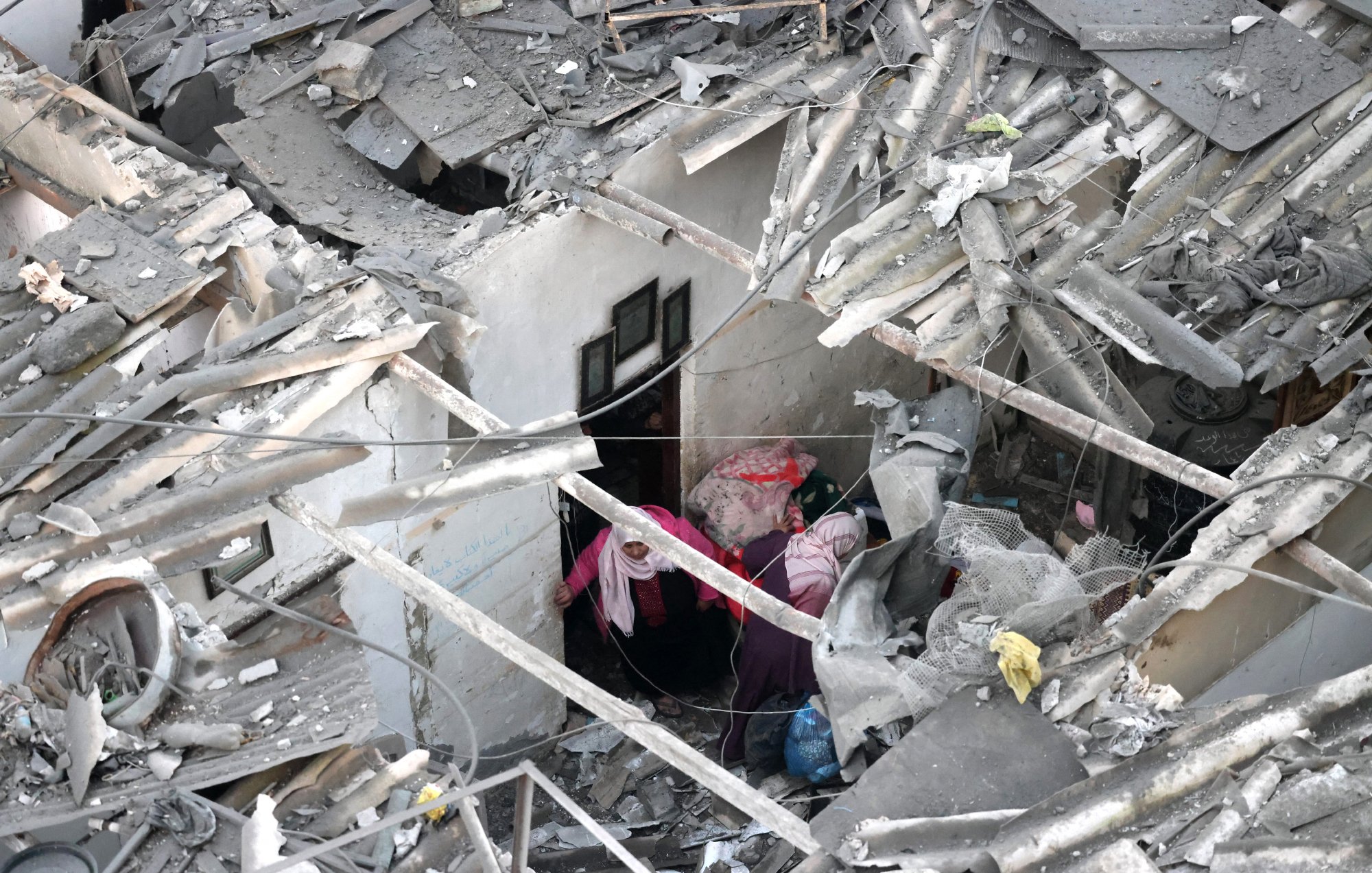 الصورة تظهر مشهداً من الدمار، حيث يوجد انخفاض في سقف أحد المنازل. debris متناثر ويرتفع الغبار في الجو، ويظهر شخصان يحاولان البحث عن شيء داخل المكان المدمر.