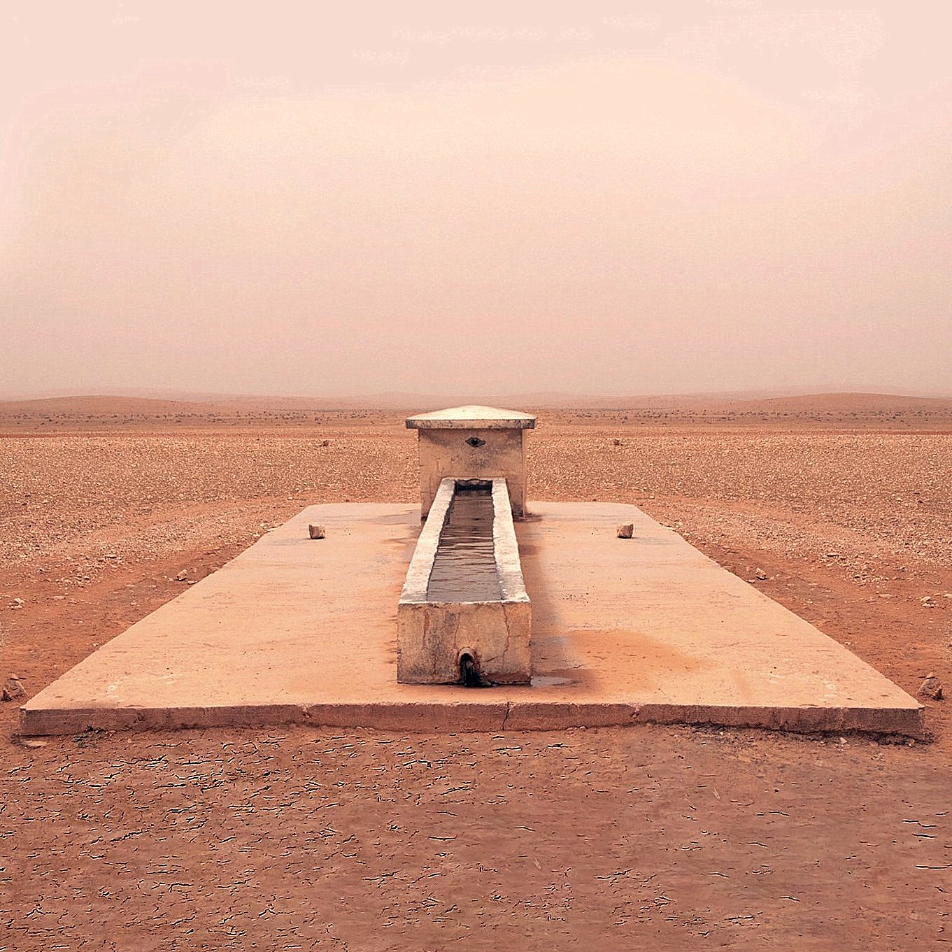 L'image montre une structure isolée située dans un paysage désertique. Au centre, il y a un long bassin en pierre, entouré par une surface plane et aride. Le sol est de couleur beige et semble sec et craquelé, tandis que l'atmosphère est légèrement brumeuse, ce qui donne une impression de chaleur et d'immensité désertique. Au loin, on aperçoit des collines ou des dunes qui se fondent avec le ciel dans une teinte douce et uniforme. L'endroit dégage une sensation de tranquillité et de solitude.